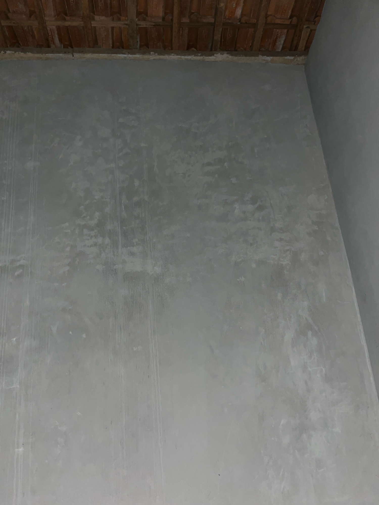 The image depicts an unfinished interior space, likely a room under construction, located in the city of Kebumen, Indonesia. The foreground is dominated by a gray, textured wall that appears to be plastered or coated with a material like concrete or stucco. This wall exhibits uneven application and some streaking, suggesting it is in the process of being finished or has been recently worked on. Vertical streaks, some darker and some lighter, are visible, possibly from tools or water.
The upper portion of the image reveals a ceiling composed of reddish-brown wooden beams and what appear to be clay roof tiles, characteristic of traditional Indonesian architecture. The junction between the ceiling and the wall is a rough, unfinished band of material.
To the right of the main wall, a corner is visible, where another gray wall meets the primary wall. This second wall is smoother and appears to be painted or finished, contrasting with the texture of the main wall. The lighting suggests an indoor environment, likely with ambient light from an unseen source.
No people, animals, or specific objects are present in the frame, emphasizing the raw, structural state of the location. The overall impression is one of an ongoing construction or renovation project.