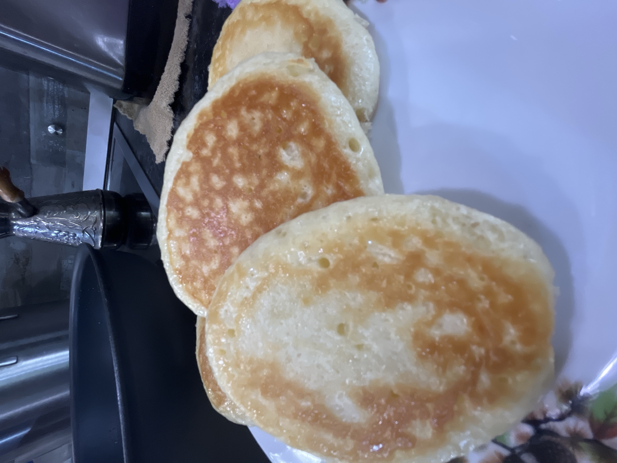 The image shows a plate of three pancakes. The pancakes are golden brown in color and appear to be freshly cooked. They are stacked on top of each other on a white plate with a floral pattern. A cooking pan is visible in the background. The image was likely taken indoors. No information from this image suggests that it was taken in Krasnodar, Russia.