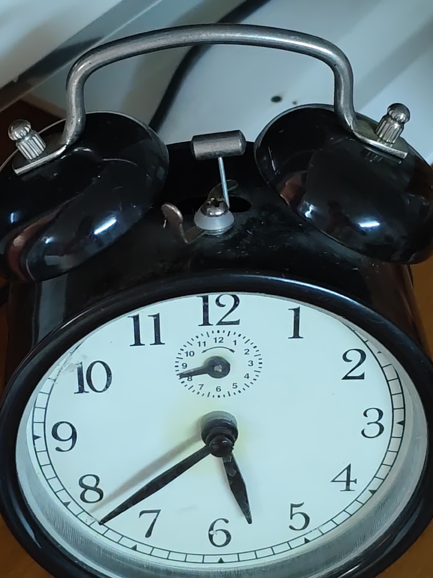 This is a close-up, high-angle, eye-level, medium shot of a black analog alarm clock with a white face. The clock is positioned on a wooden surface. The time displayed on the clock is approximately 8:59, with the minute hand pointing just before the 9 and the hour hand pointing just before the 9. The clock has a metallic silver handle on top and two bells on either side. The face of the clock has large black numbers for the hours and smaller markings for minutes and seconds. The alarm clock is the sole subject in the image. The background is blurred and appears to be a white surface. This image was not taken in Olginskaya, Russia; the provided image does not contain any information about the location.