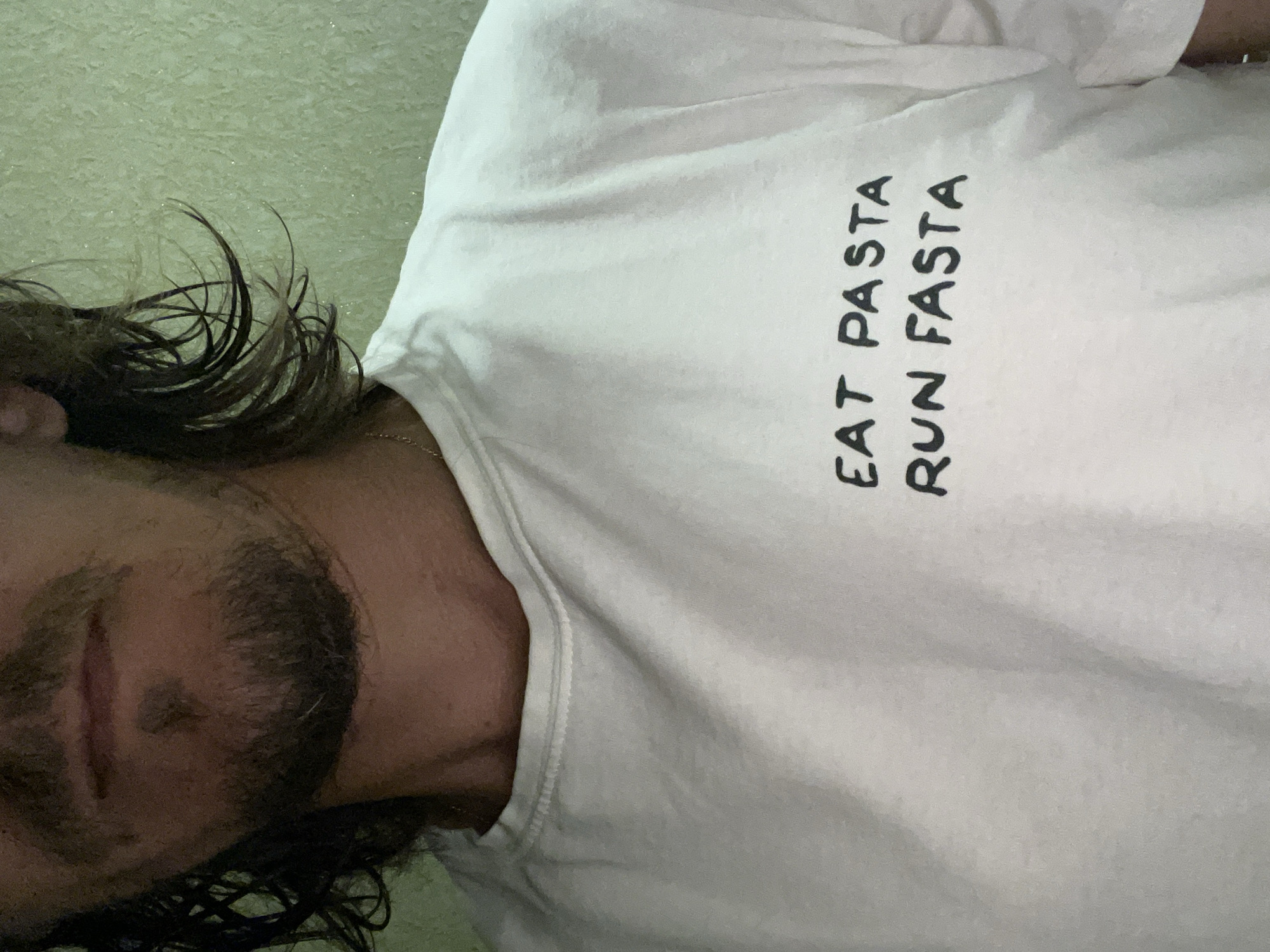 The media file is a close-up, rotated, vertical shot of a person wearing a white t-shirt. The t-shirt has black text printed on it that reads "EAT PASTA RUN FASTA". The person's face is partially visible, showing a section of their eye and mouth, along with their hair and beard. The background appears to be a textured, light green wall. The lighting is soft, and it is unclear what time of day it is or what the overall weather conditions are. There are no other people or objects visible in the frame. The focus is on the text on the t-shirt and the person's face.