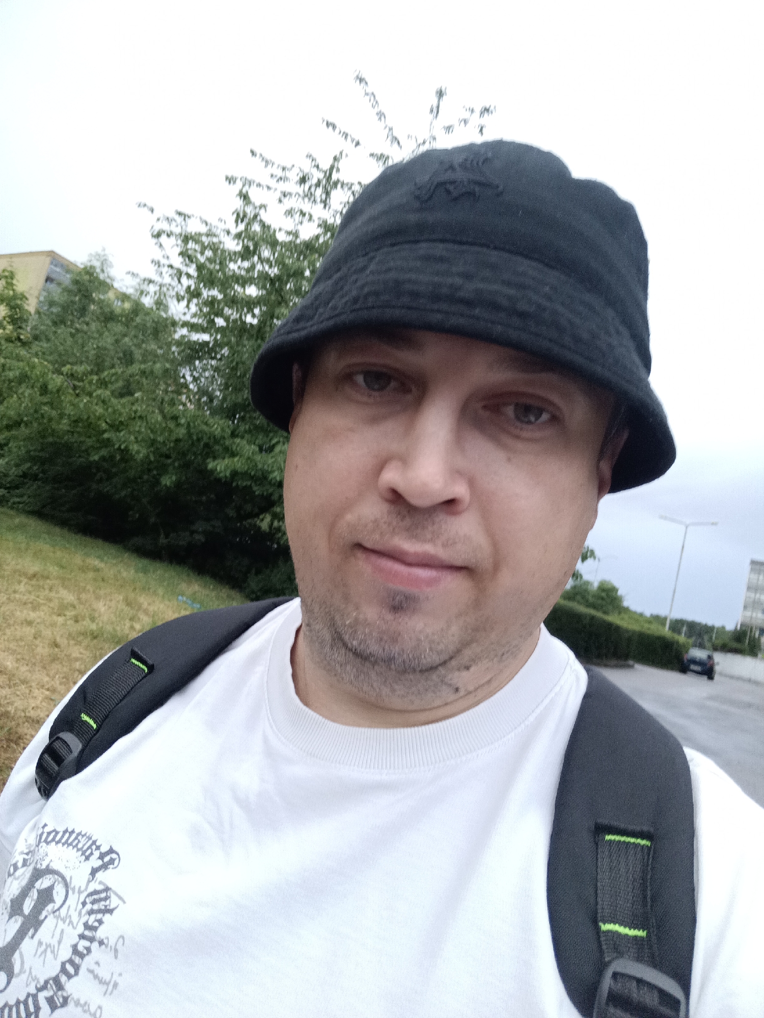 A man wearing a black bucket hat and a white t-shirt with a backpack is taking a selfie outdoors. He has short, brown hair and stubble. The background shows green trees, a grassy slope, a road with a car, and buildings. The sky is overcast. The scene appears to be in a suburban or park area. There is no indication of the city of Prague, Czechia in the image.