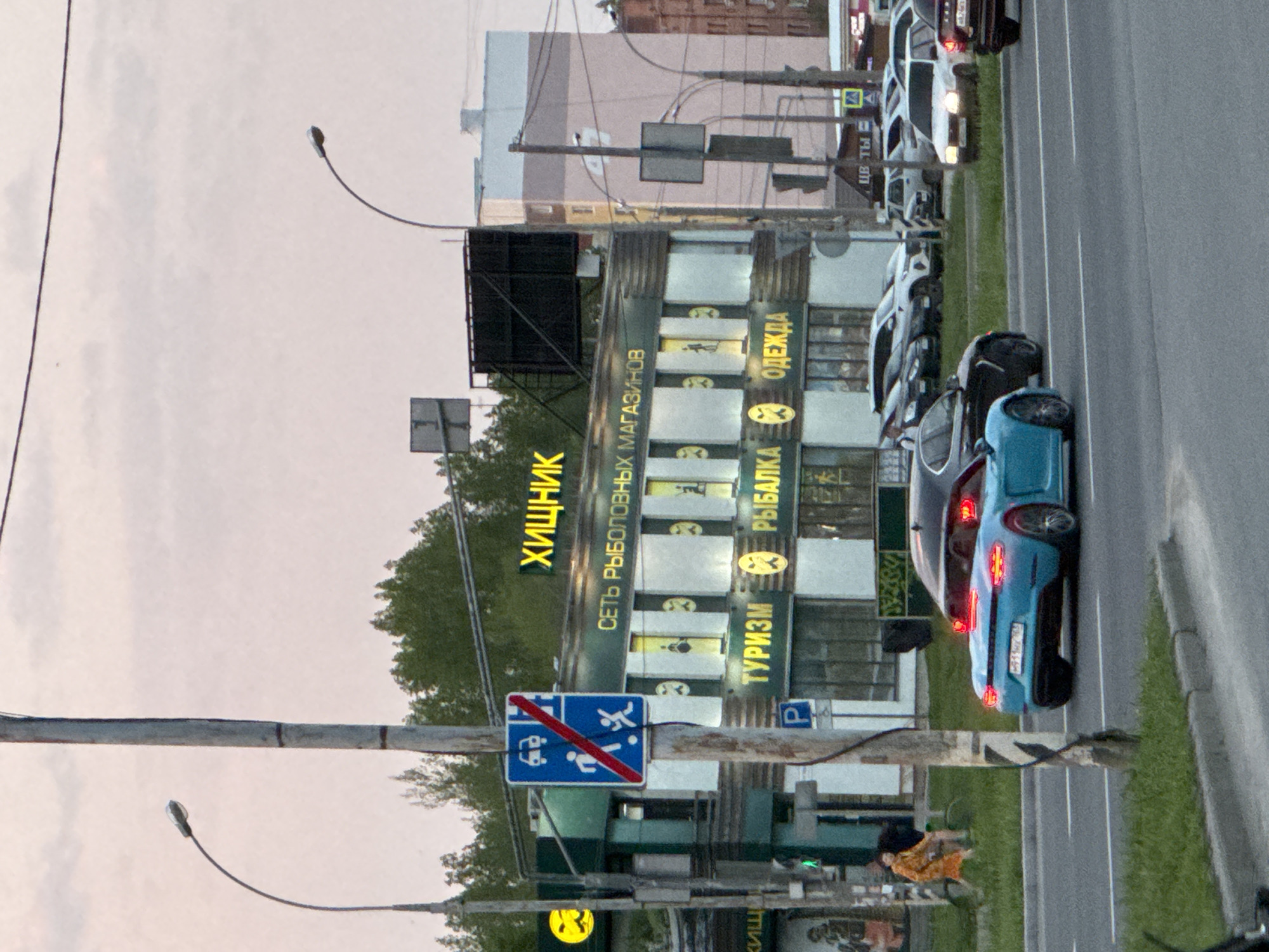 The image shows a city street with several parked cars, a sidewalk, and a building on the right. The building appears to be a store, with signs written in a language other than English (likely Russian), advertising goods for tourism, fishing, and clothing. A street sign is visible, indicating that certain forms of transportation are restricted (likely pedestrians and vehicles). The street scene is urban, with light poles and electrical lines visible. There is also a person near the sidewalk in an orange jacket. The sky appears overcast or dusky.