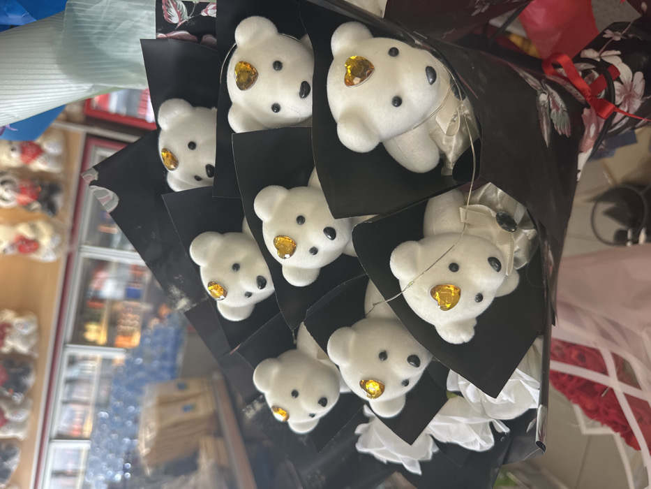 A close-up, slightly angled photograph depicts a bouquet composed of approximately nine small, white plush teddy bears, presented in an indoor retail setting in Torbalı, Turkey. Each teddy bear features black bead eyes, a black stitched nose, and a prominent golden heart-shaped jewel affixed to its forehead. Some of the bears are adorned with small, light-colored bows around their necks, and delicate fairy lights are interwoven among them.
The bears are individually nested within black, conical paper wrappers, interspersed with white artificial roses. The entire arrangement is encased in a larger black floral wrapping paper, with a section of patterned paper and a red ribbon visible at the lower right.
The background is intentionally blurred, revealing elements of a store interior. Visible are several display shelves stocked with various packaged items, along with refrigerated display units containing beverages. In the upper background, additional stuffed animals, consistent with teddy bears, are visible on higher shelves. No individuals are present, and no specific actions or interactions are depicted within the frame.