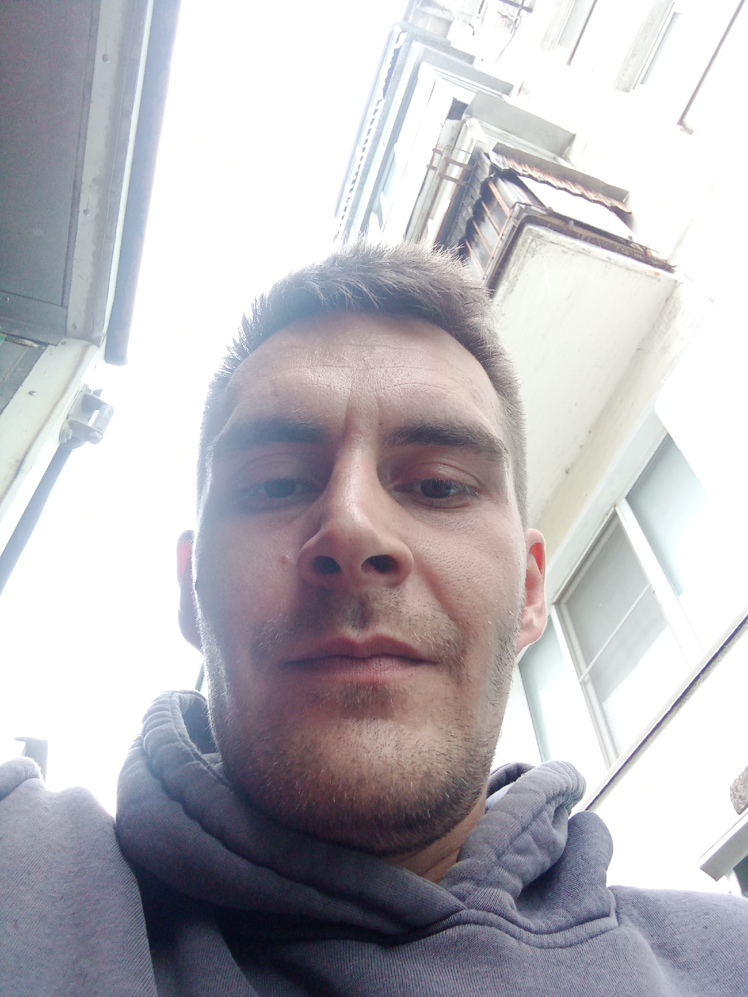 A low-angle, head-and-shoulders selfie captures a man looking directly at the camera. He appears to be in his 20s or 30s, with short, dark hair and light stubble on his face. He has a neutral, somewhat serious expression. He is wearing a grey hooded sweatshirt, with the hood visible around his neck.
The background is highly overexposed and bright, suggesting a clear, sunny daytime outdoors. Above and behind him, parts of urban buildings are visible from a worm's-eye view. On the left, a light-colored structure with vertical lines and a pipe-like fixture can be seen. On the right, a multi-story building features balconies or ledges with visible details, including what looks like a small, slightly damaged roof or cover over one of them, and windows reflecting the bright light. The architecture appears functional, typical of older urban residential or commercial structures in Chaikovsky, Russia. There is no visible text in the image.