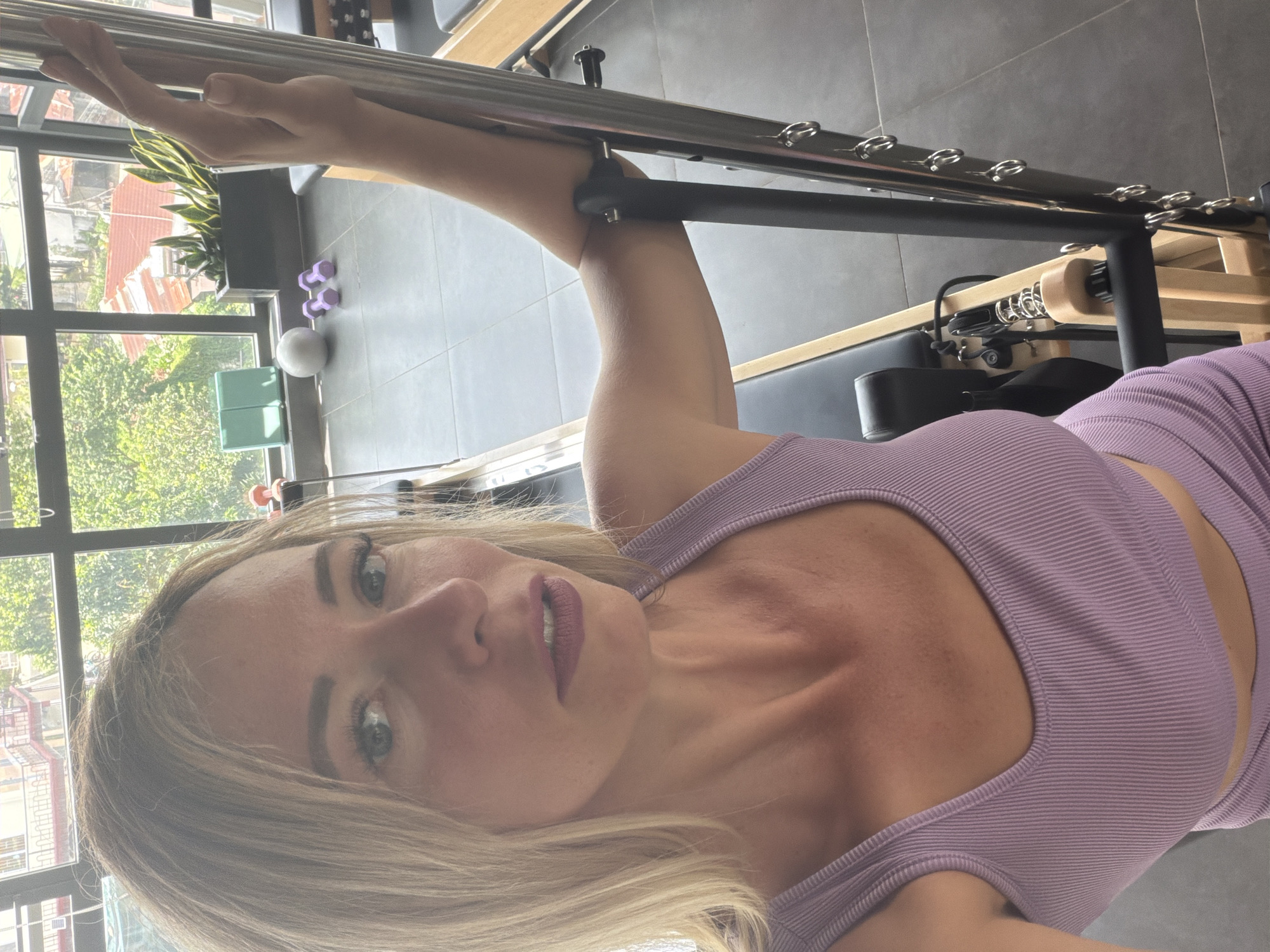 A woman with blonde hair and light skin is taking a selfie in a gym. She is wearing a purple, ribbed tank top and her lips are painted with a dark lipstick. Her left arm is extended upwards, grasping a metal bar. The background shows a Pilates reformer machine, with its wooden frame and padded seat. In the background, through a large window, there is a view of buildings and lush green trees, suggesting an outdoor scene with daytime light. Several small purple dumbbells and a white exercise ball are visible on the floor. The overall impression is of a woman engaged in a fitness activity in a well-lit studio.