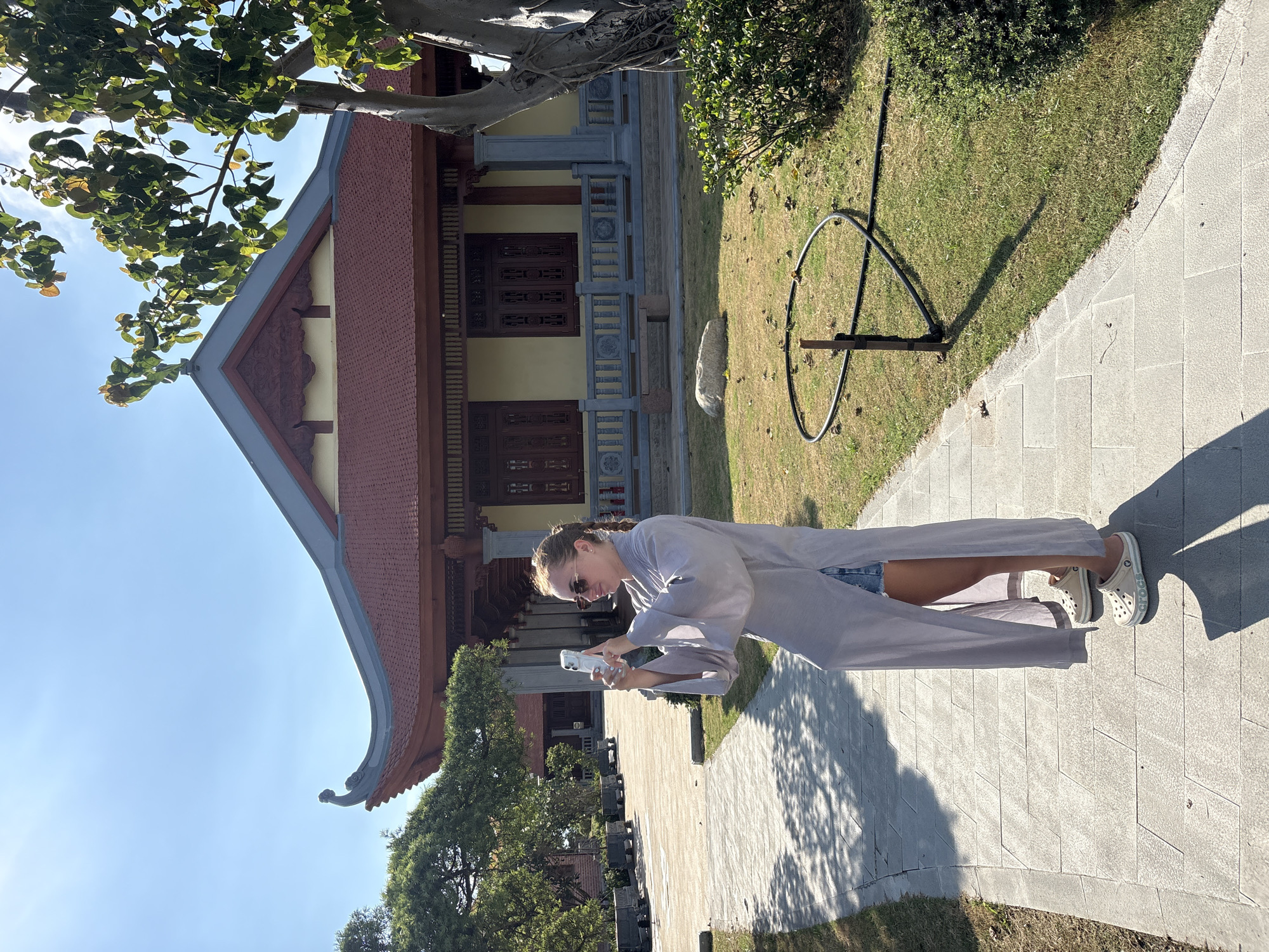 A woman stands on a paved walkway in the city of Tay Nha Trang Ward, Vietnam, holding and looking at a white smartphone. She is wearing a long, light-colored robe or overcoat, denim shorts, and cream-colored Crocs shoes. Her hair is pulled back. Behind her is a traditional Vietnamese building with a tiered roof, a yellow facade, and dark brown wooden details. To the right of the building, there is a patch of green grass with a circular metal sculpture. Trees and a clear blue sky are also visible.