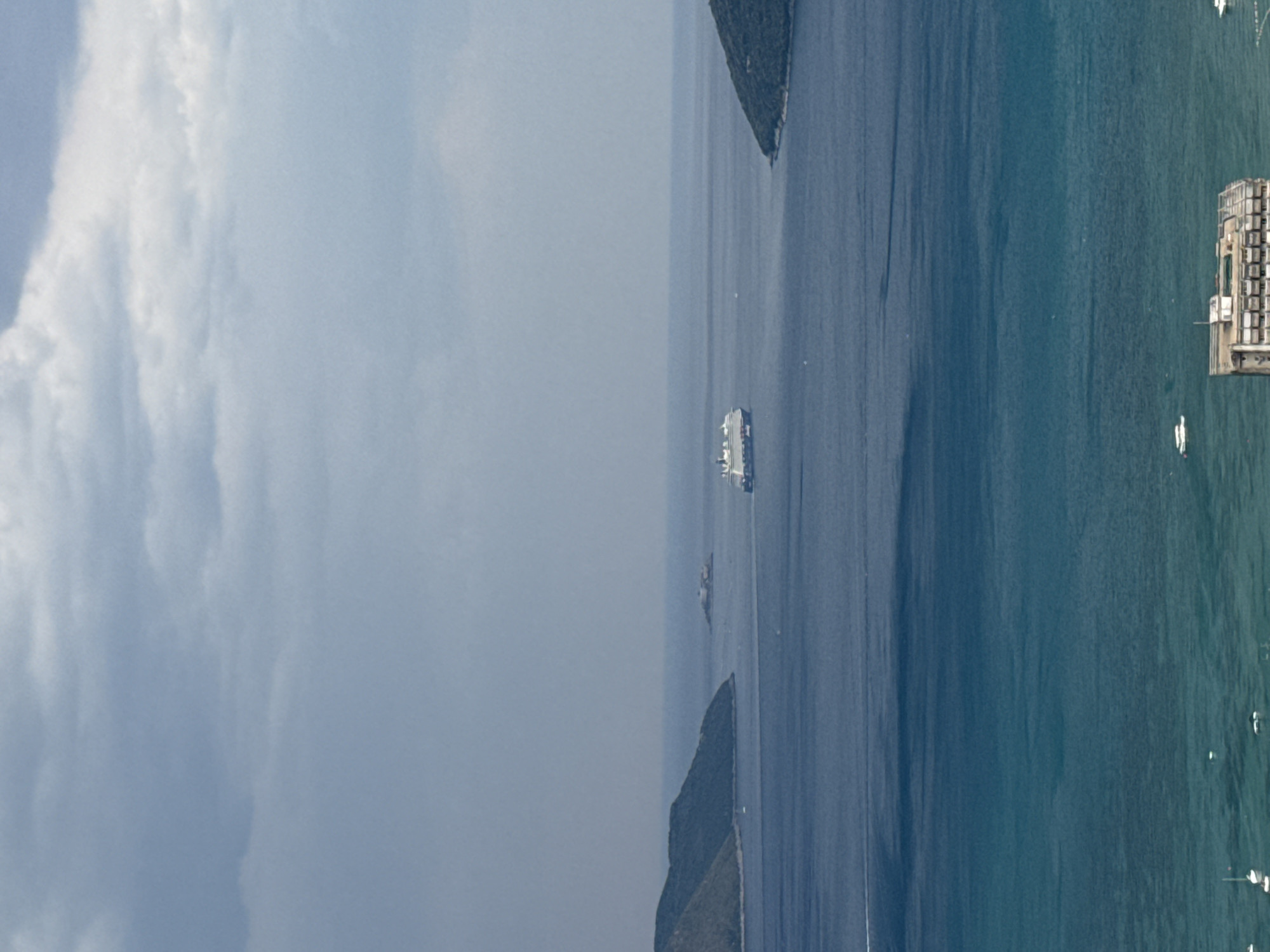 This is an aerial photograph taken from a high vantage point, likely a drone or aircraft. The image depicts a coastal scene with a body of water, possibly a bay or sea, meeting a landmass covered in lush green vegetation. Several boats are visible scattered across the water, suggesting a leisurely or commercial maritime activity. In the distance, a large white vessel, possibly a ferry or cruise ship, is navigating the water. On the right side of the frame, a complex of buildings, appearing to be industrial or resort structures, stands on the coastline, with more boats docked nearby. The water transitions in color from a deep blue to a lighter, more greenish hue closer to the shore, indicating depth variations or changes in water composition. The sky is partly cloudy, with soft, diffused light suggesting it is daytime, possibly late morning or afternoon. The scene conveys a sense of calm and expanse, with the natural beauty of the coastline juxtaposed with man-made structures and vessels. The overall impression is of a serene, active coastal environment.