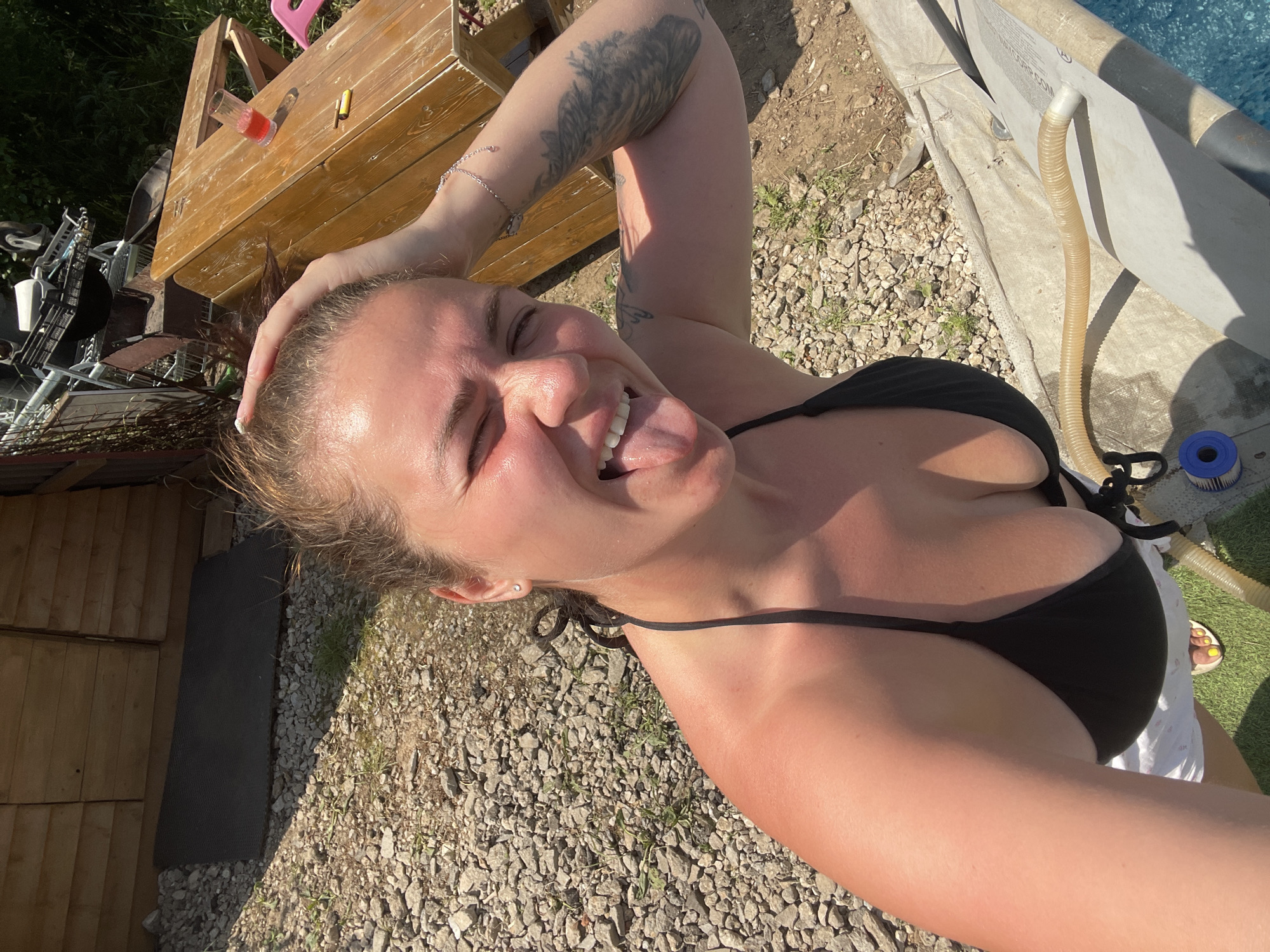A woman is pictured outdoors, in what appears to be a backyard or recreational area. She is wearing a black bikini top and white shorts, and her hair is pulled back. She has a playful expression, with her tongue sticking out and her hand on her head. The background shows a wooden structure, a picnic table with a drink and a cigarette, and a pool with a hose. The ground is covered in gravel and some grass. There is also some industrial-looking equipment and a shopping cart in the background. The lighting suggests it is a sunny day. The location is described as Мосметаллоконструкция Онт, Russia.