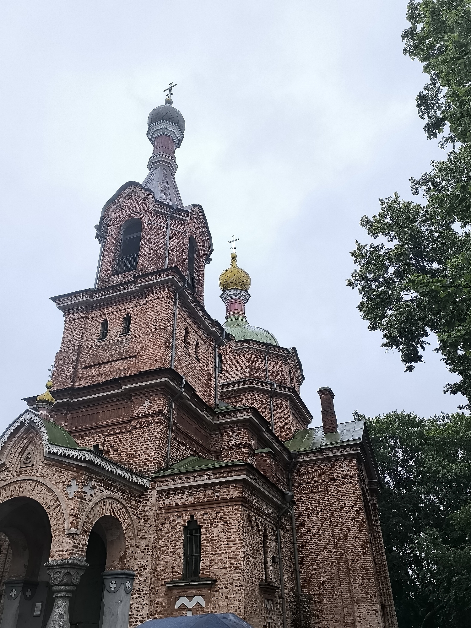 The image shows a brick church with multiple domes and a bell tower, situated in Kuldīga, Latvia. The architecture is predominantly red brick, with some green-domed roofs and golden accents on the highest dome. There are visible crosses atop the domes. The sky is overcast, and the upper portion of a large green tree is visible on the right side of the frame, extending into the sky. The foreground includes the arched entrance of the church. The perspective is from a low angle, looking up at the church's structure.