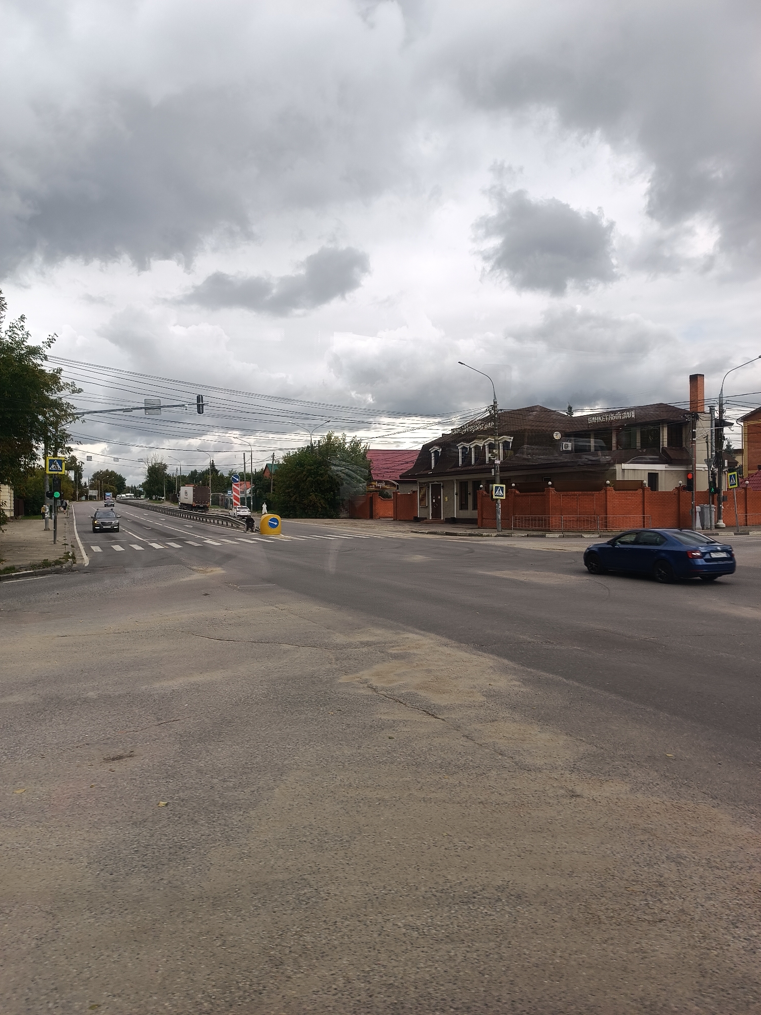 The image captures a wide, paved road in what appears to be a suburban area, possibly Zhilino-1, Russia, under a cloudy sky. A dark blue sedan is driving on the right side of the road, turning. In the background, on the right side of the road, is a large building with a brown roof, featuring a brick chimney and multiple windows. The building has signage that reads "БАНКЕТНЫЙ ЗАЛ" (Banquet Hall). A brick fence encloses the property. On the left side of the road, there is a pedestrian crossing with a traffic light showing a green signal. A dark-colored car is stopped at the crossing, and a few people are visible on the sidewalk. Numerous overhead wires are strung across the road. The road surface shows signs of wear, with patches of lighter-colored material.