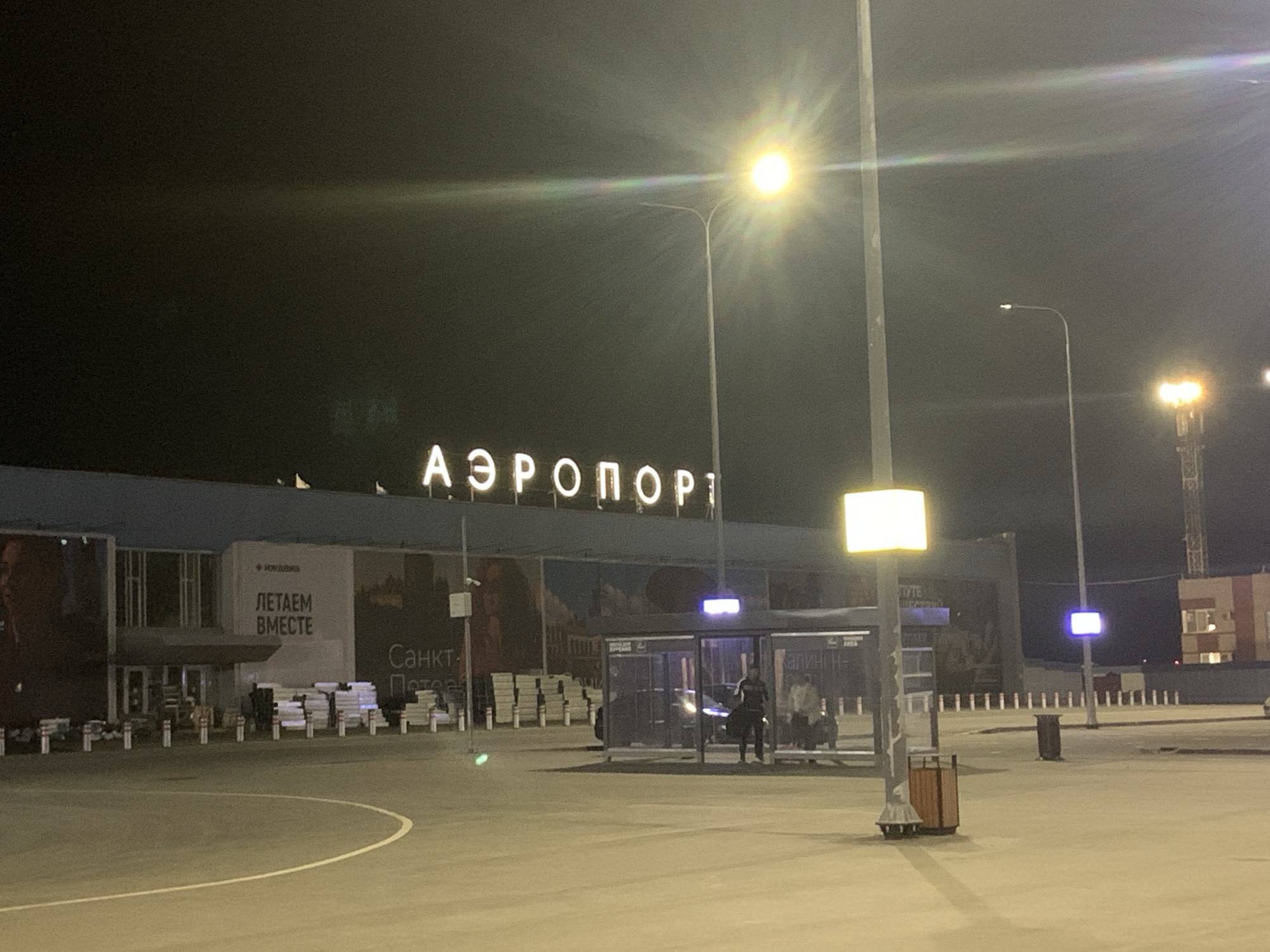 The image depicts the exterior of an airport at night, with bright lights illuminating the facade and surrounding area. A large sign above the building reads "АЭРОПОРТ" (AEROPORT), indicating it is an airport. To the left of the sign, a smaller banner displays text that translates to "WE FLY TOGETHER." There are several streetlights and illuminated signs visible, some with a strong glare. In the foreground, there is a paved area with a curved white line, resembling a road or parking lot. A bus stop shelter with people inside and waiting is visible in the middle ground. A trash receptacle is situated near a lamppost. The overall scene suggests a functional, well-lit airport environment during nighttime operations. The specific location is the Izhevsk Airport (IJK) in Izhevsk, Russia.