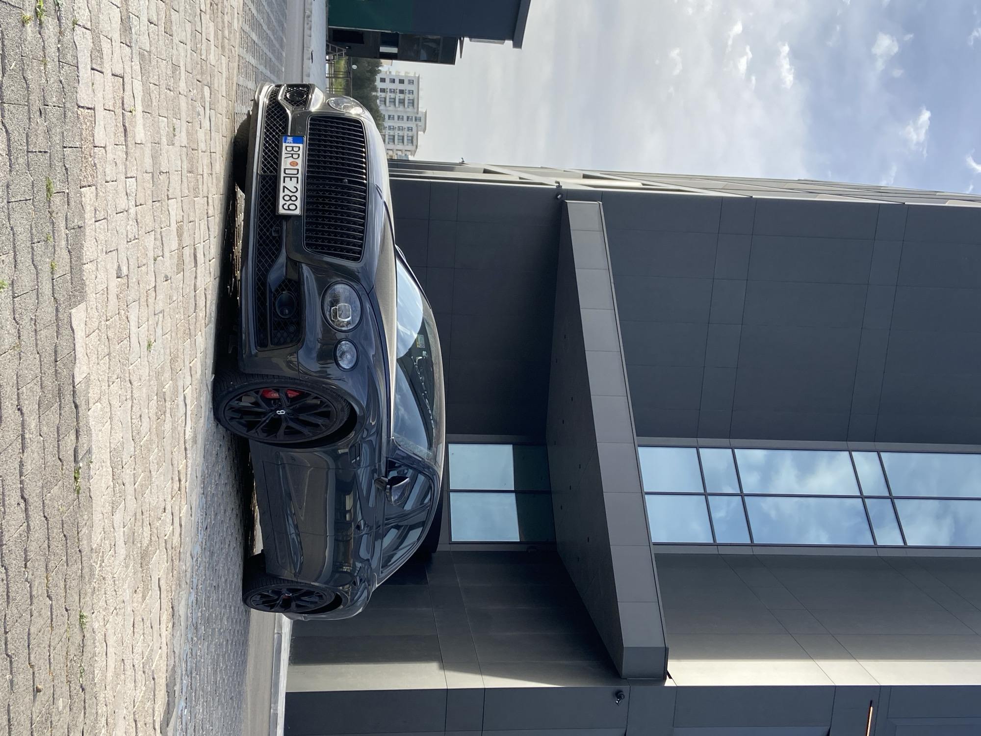 A black Bentley is parked on a cobblestone street next to a modern building with large windows. The car has black wheels and red brake calipers. The license plate is visible, "BR DE 289". In the background, another building is visible, and the sky is partly cloudy. The location is Bar, Montenegro, as indicated by the license plate. The car is the primary subject, and the environment suggests an urban setting.