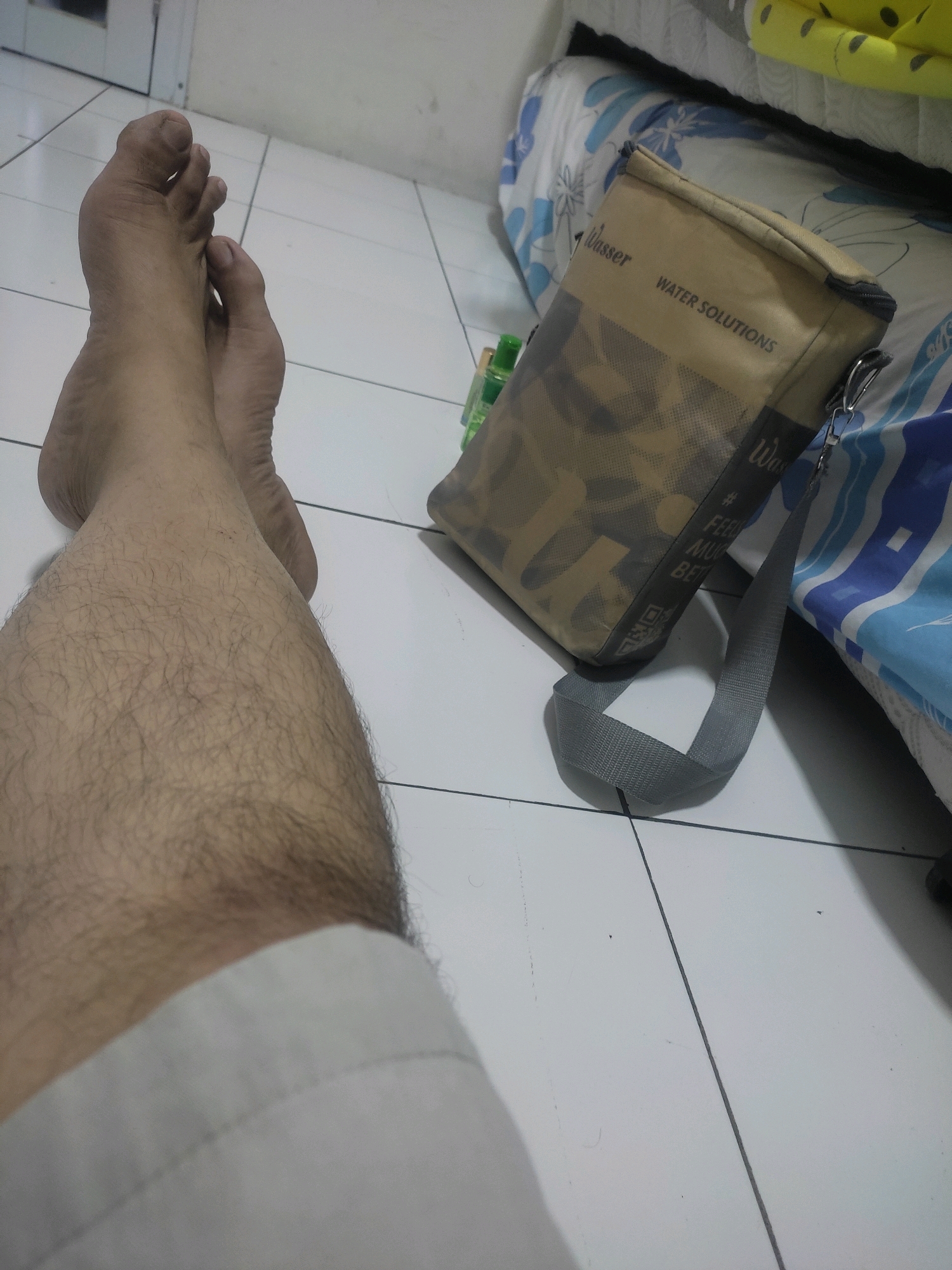 The image depicts a person's legs and feet resting on a tiled floor, with a patterned bag and a bed in the background. The person is wearing light-colored shorts. The bag is a light brown color with a camouflage pattern and text that reads "Wasser WATER SOLUTIONS". A grey strap is attached to the bag. The bed has a blue and white patterned comforter. The overall scene suggests an indoor setting, possibly a bedroom or living space. The floor is white tile with dark grout lines. The lighting appears to be artificial, casting shadows on the floor. There are no discernible landmarks to place the image specifically within Bogor, Indonesia.