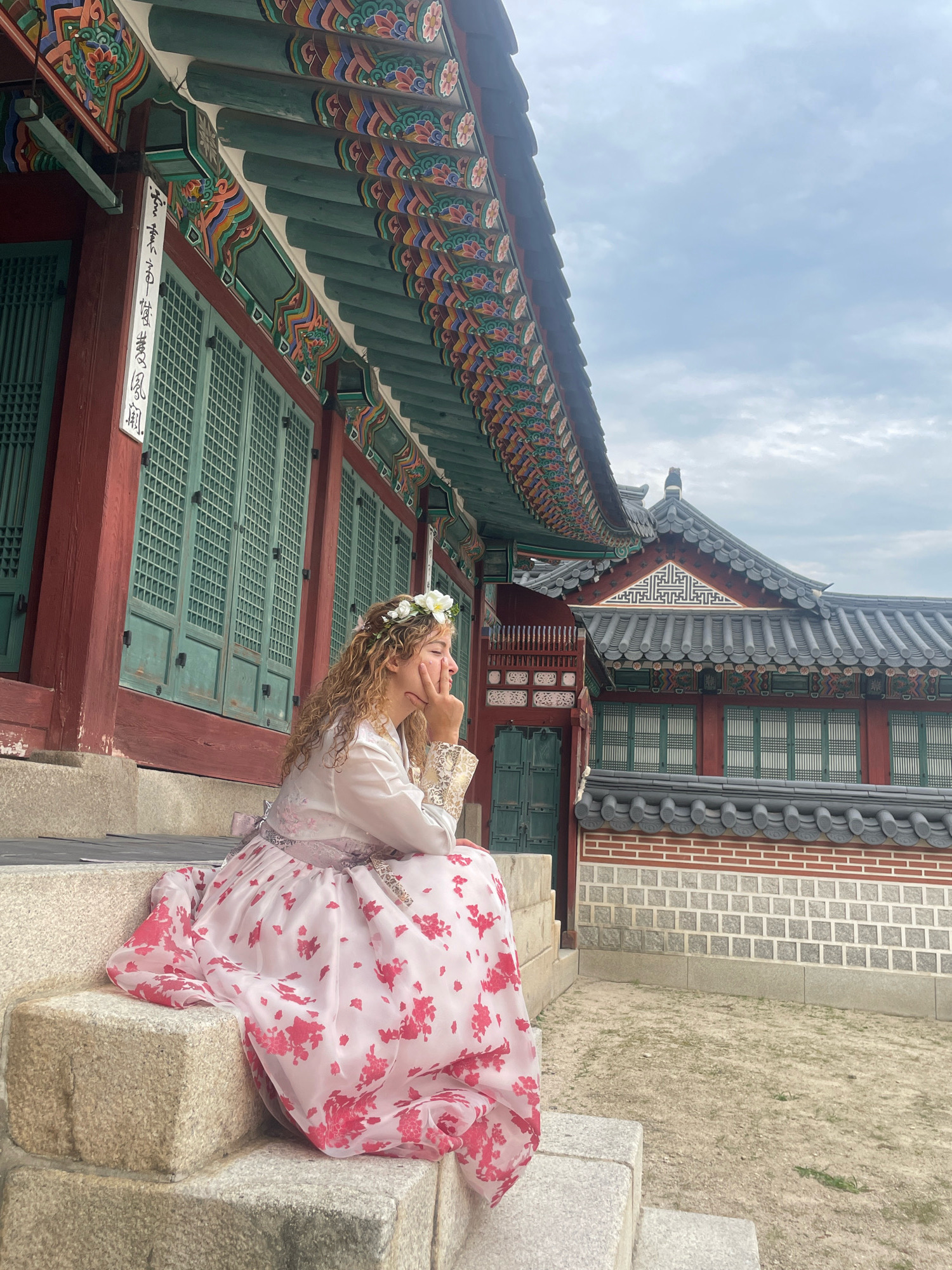 In Seoul, South Korea, a young woman is captured in a full-body shot, seated on the stone steps of a traditional Korean palace. She wears a vibrant pink and white hanbok adorned with floral patterns, complemented by a delicate white flower crown. Her posture is relaxed as she gazes thoughtfully to the side, her hand resting on her chin. Behind her, the ornate architecture of the palace unfolds, characterized by its distinctive green tiled roofs, colorful decorative eaves, and wooden window lattices. The scene is set against a cloudy sky, hinting at a temperate day. The overall impression is one of serene contemplation amidst historical grandeur.