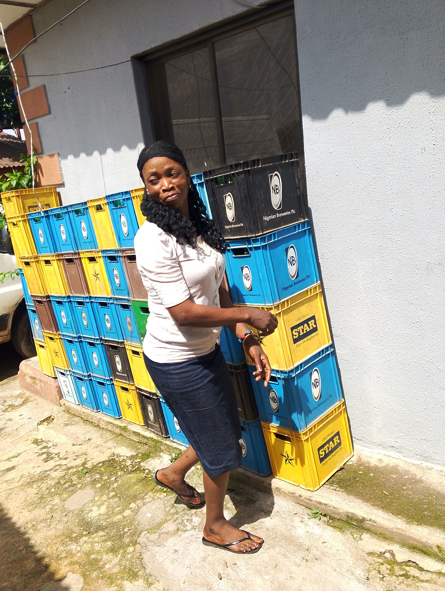A Black woman with dark braided hair and a headscarf stands in front of a wall in Jos, Nigeria. She is wearing a light-colored short-sleeved shirt and a dark skirt. She is standing next to a large stack of beer crates, predominantly blue and yellow, with some brown crates interspersed. The crates are branded with "NB Nigerian Breweries Plc" and "STAR Cold Filtered". The ground in front of the crates is uneven concrete with patches of moss and dirt. Sunlight casts a distinct shadow on the wall behind the crates. A small portion of a white vehicle is visible to the left of the crates.