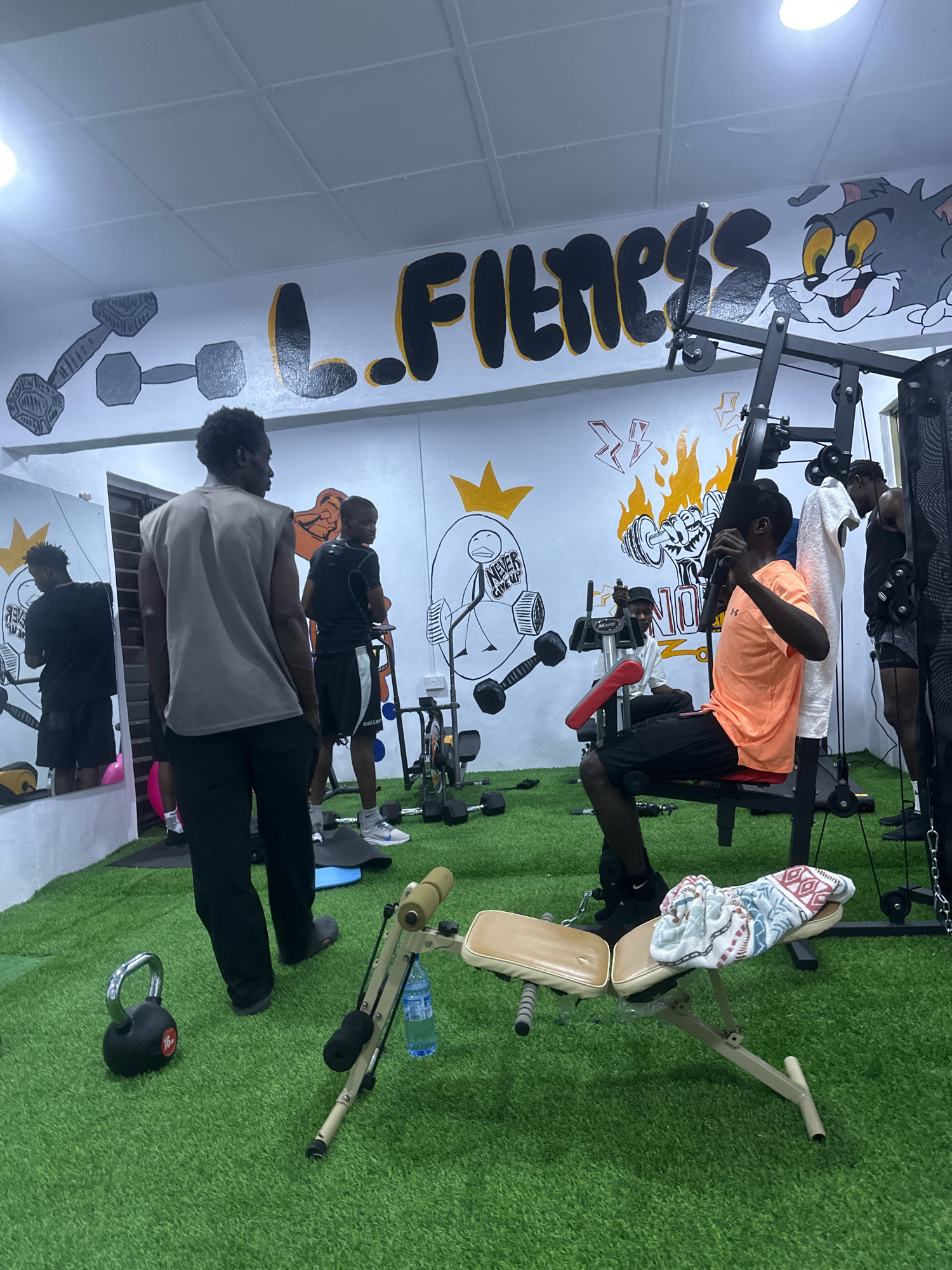 In Badagry, Nigeria, a gym interior is depicted with several individuals engaged in workout activities. The floor is covered with artificial green turf, and the walls are adorned with vibrant murals. One mural features a large, stylized inscription of "L. Fitness" in bold black and yellow lettering, accompanied by cartoonish drawings of a dumbbell and the character Tom from Tom and Jerry. Another mural depicts a figure lifting a dumbbell with flames surrounding it.
Several men are present in the gym. In the foreground, a man wearing a gray sleeveless shirt and black pants stands facing away from the camera, with his hands in his pockets. To his right, another man in an orange t-shirt and black shorts is seated on a weight machine, actively using it. Behind them, a man in a black t-shirt and shorts is also on a weight machine. Another individual in a black t-shirt and shorts is visible to the far right, appearing to be preparing for a workout. In the background, a man in a black shirt and shorts is reflected in a large mirror on the left side of the room.
Various gym equipment is scattered throughout the space, including weight machines, dumbbells, and a kettlebell marked "16kg." A portable weight bench with a patterned towel draped over it is positioned in the mid-ground. A water bottle is attached to a piece of equipment. The lighting in the gym appears to be from overhead fluorescent fixtures.