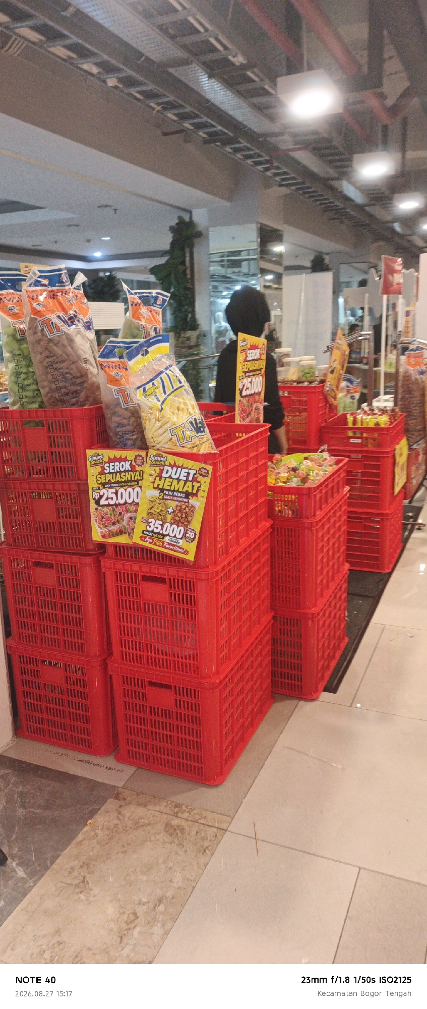 The image depicts a retail environment, likely a supermarket or a snack shop, in Bogor, Indonesia. In the foreground, several stacked red plastic crates are prominently displayed, containing various packaged snacks. One crate features a yellow sign with red text that reads "SEROK SEPUASNYA!" and "DUET HEMAT," indicating a promotion or sale. Bags of snacks, including what appear to be potato sticks and other savory items, are visible within the crates.
In the middle ground, a person wearing a dark head covering and a dark shirt is partially visible, facing away from the camera and towards the display of snacks. Behind the person, shelves and more products are visible, suggesting a larger retail space. The background is blurred but shows overhead lighting fixtures and ceiling structures typical of an indoor commercial setting.
The floor appears to be tiled with different patterns, with some lighter tiles and some with a marbled effect. The overall scene suggests a busy shopping area where snacks are being advertised and sold. The timestamp at the bottom of the image indicates it was taken on August 27, 2026, at 15:17, with camera settings of 23mm f/1.8 1/50s ISO2125, and the location is specified as Kecamatan Bogor Tengah, Bogor, Indonesia.