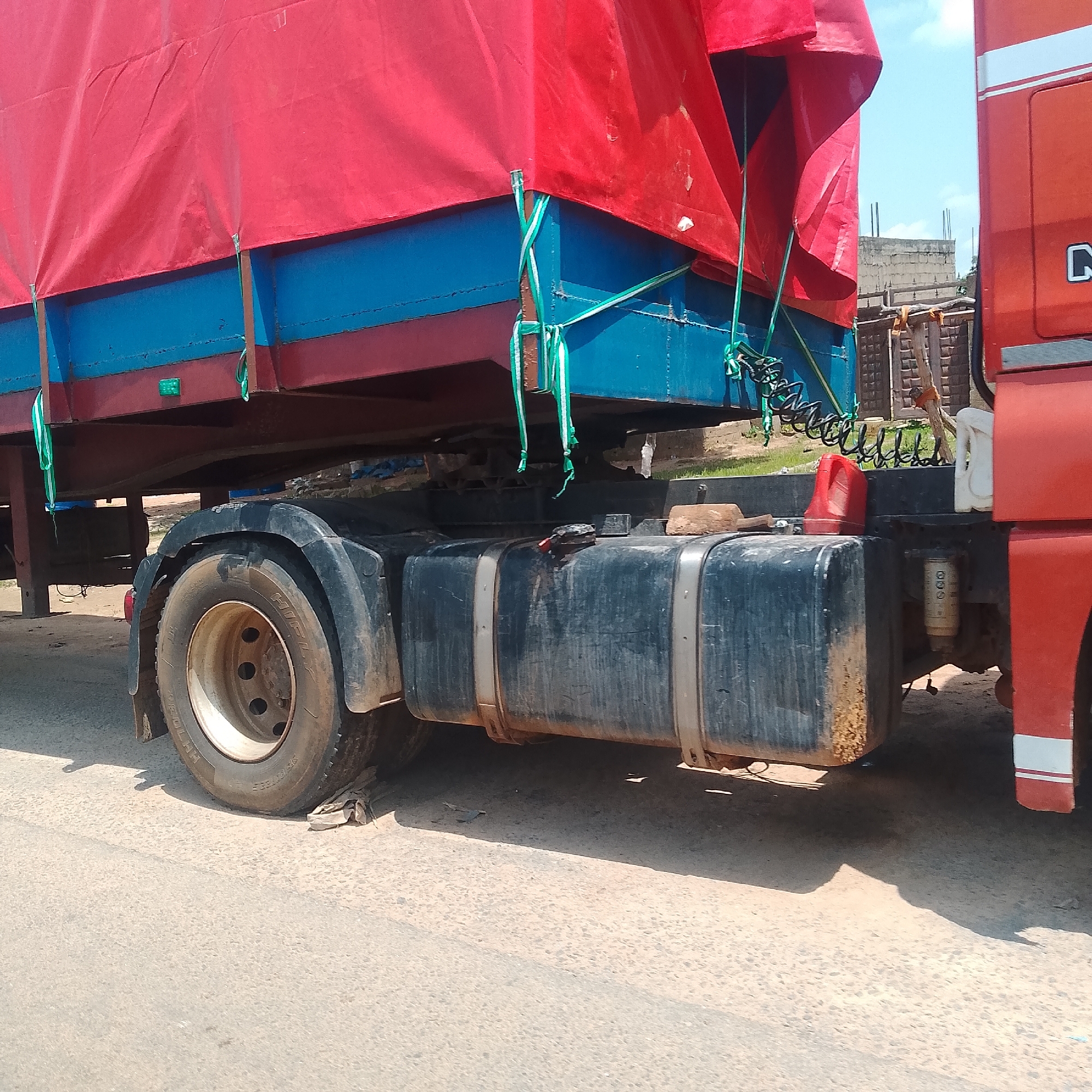 The image shows the side view of a red and blue truck trailer parked on a paved road in Jalingo, Nigeria. The trailer's cargo is covered with a large red tarp, secured with green and white straps. The truck's large black fuel tank is visible below the trailer. The scene appears to be in an urban or semi-urban area, with a building and some construction materials visible in the background. The lighting suggests it is daytime, with a clear sky. There are no people visible in the image. The truck is stationary, and the overall impression is one of a commercial vehicle either parked or stopped during transit.