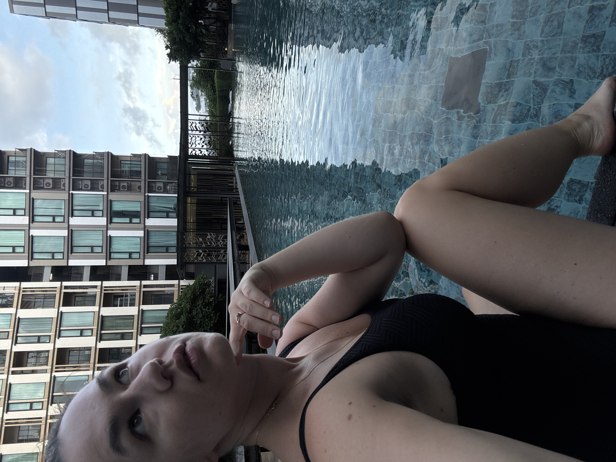 A woman in a black swimsuit sits by a swimming pool with buildings in the background. She is looking up and to her left with her chin resting on her hand. The water in the pool reflects the buildings and the sky. The scene is likely set in Ratsada, Thailand, given the architecture and tropical foliage.