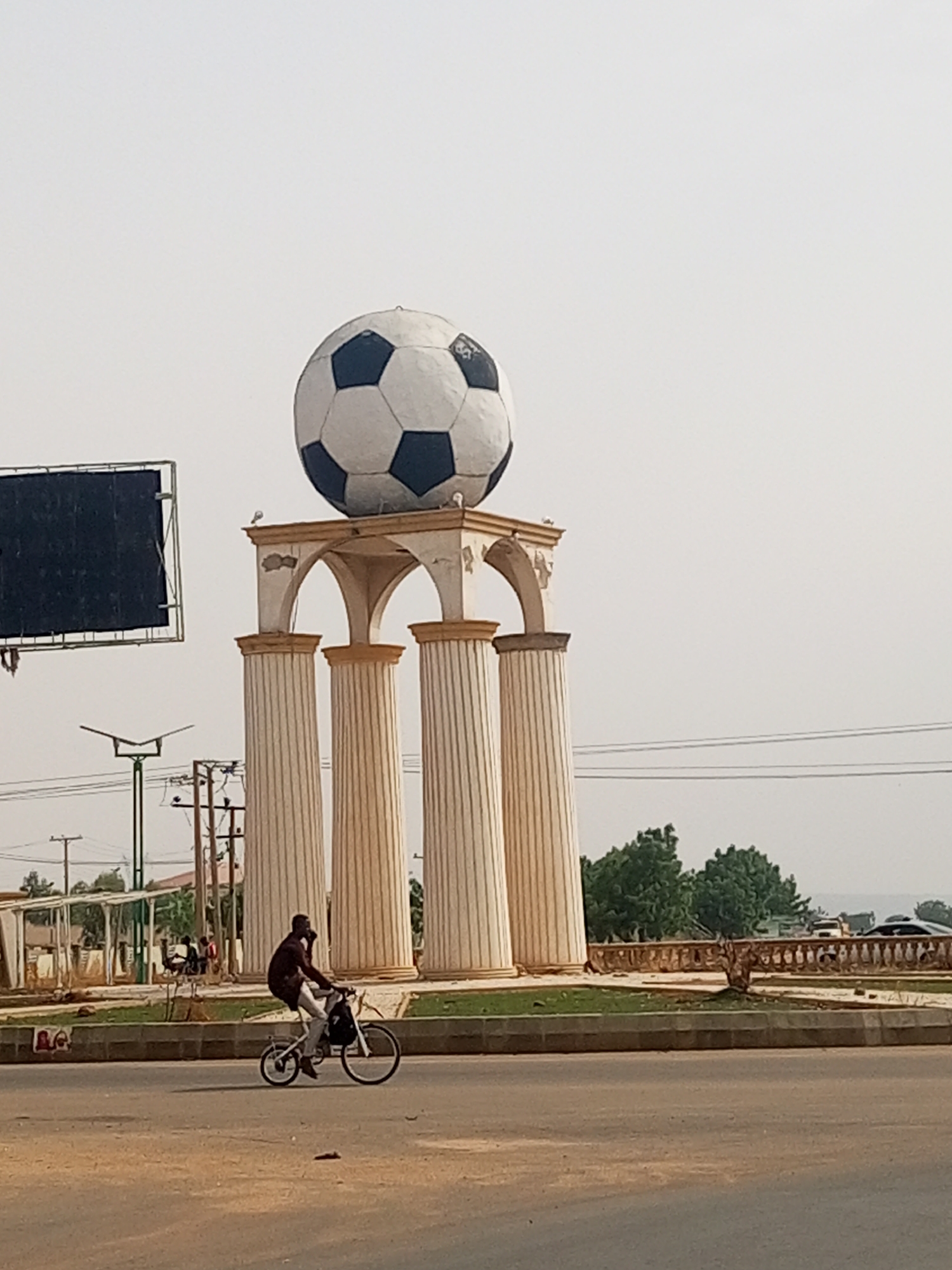 The image displays a prominent football monument in an urban setting during daytime, likely a roundabout in Gombe, Nigeria. At the center, a large, black and white soccer ball is mounted on a classical-style pedestal, featuring four fluted columns and decorative arches painted in light beige and tan tones. Small birds are perched on the monument's structure.
In the foreground, a man rides a white bicycle from left to right across the road. He is dressed in dark clothing and light-colored trousers, with what appears to be a bag attached to his bicycle or carried on his back. The road surface consists of a light-colored, possibly dusty, asphalt.
To the left, a large, dark, rectangular billboard frame is visible, appearing blank. The background includes utility poles with power lines, green trees, and distant hazy structures under a bright, somewhat overcast or hazy sky. The scene suggests a busy, warm, and dry environment. No discernible text is visible within the image.