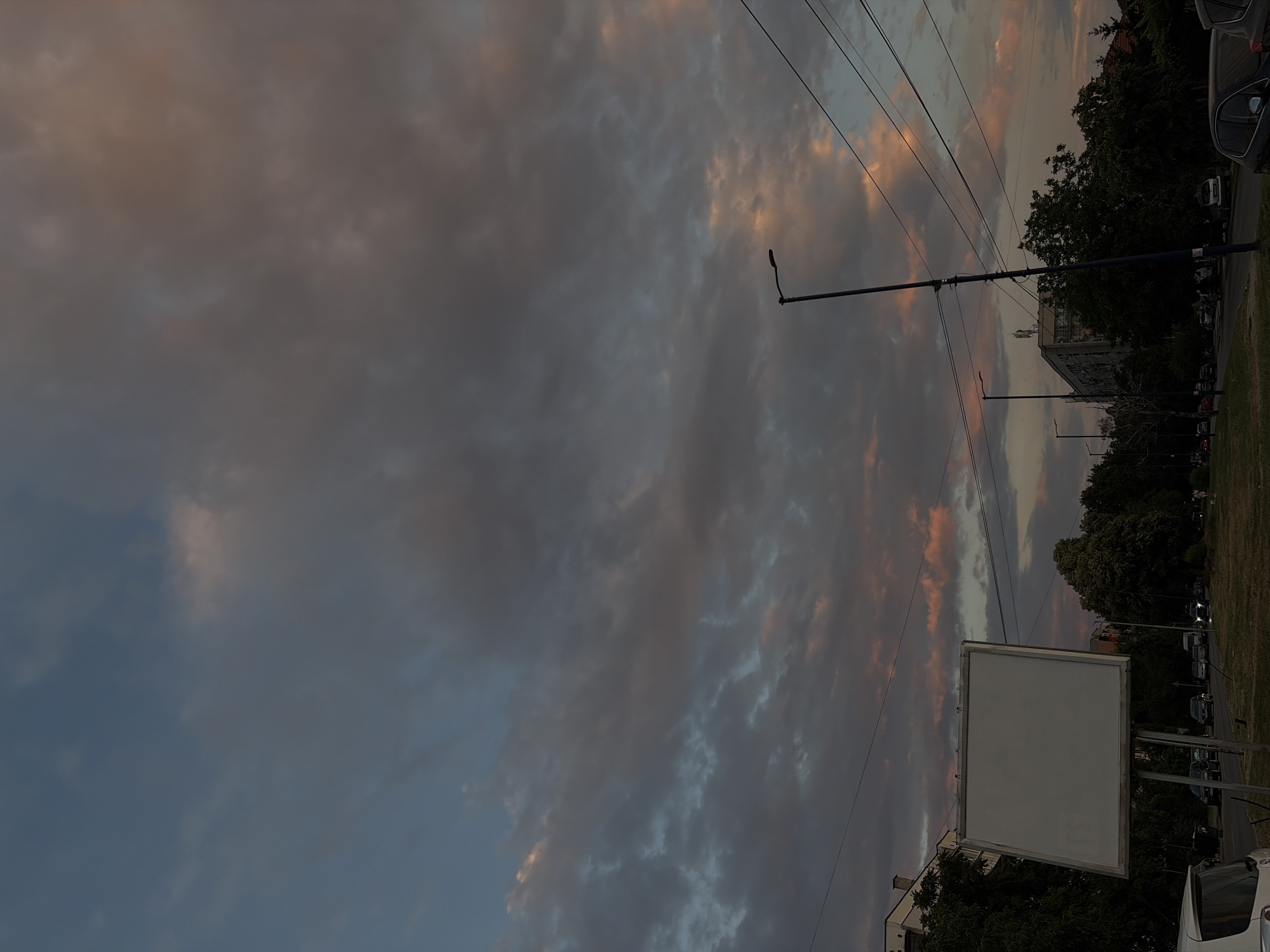 The image depicts an urban scene at dusk. The sky is filled with dramatic clouds, tinged with hues of pink and orange, contrasting with darker gray and blue tones. In the foreground, a large, blank white billboard stands prominently on a metal frame. To the right of the billboard, a tall street lamp with a single light fixture is visible, along with a network of power lines crisscrossing the frame. The middle ground features a tree-lined street with cars parked and driving, indicating a roadway. Buildings, some appearing to be apartment complexes, are visible behind the trees and street lights. In the bottom left corner, the rear of a white vehicle is partially visible. The overall atmosphere is moody and somewhat dramatic, characteristic of a sunset or twilight in an urban environment.