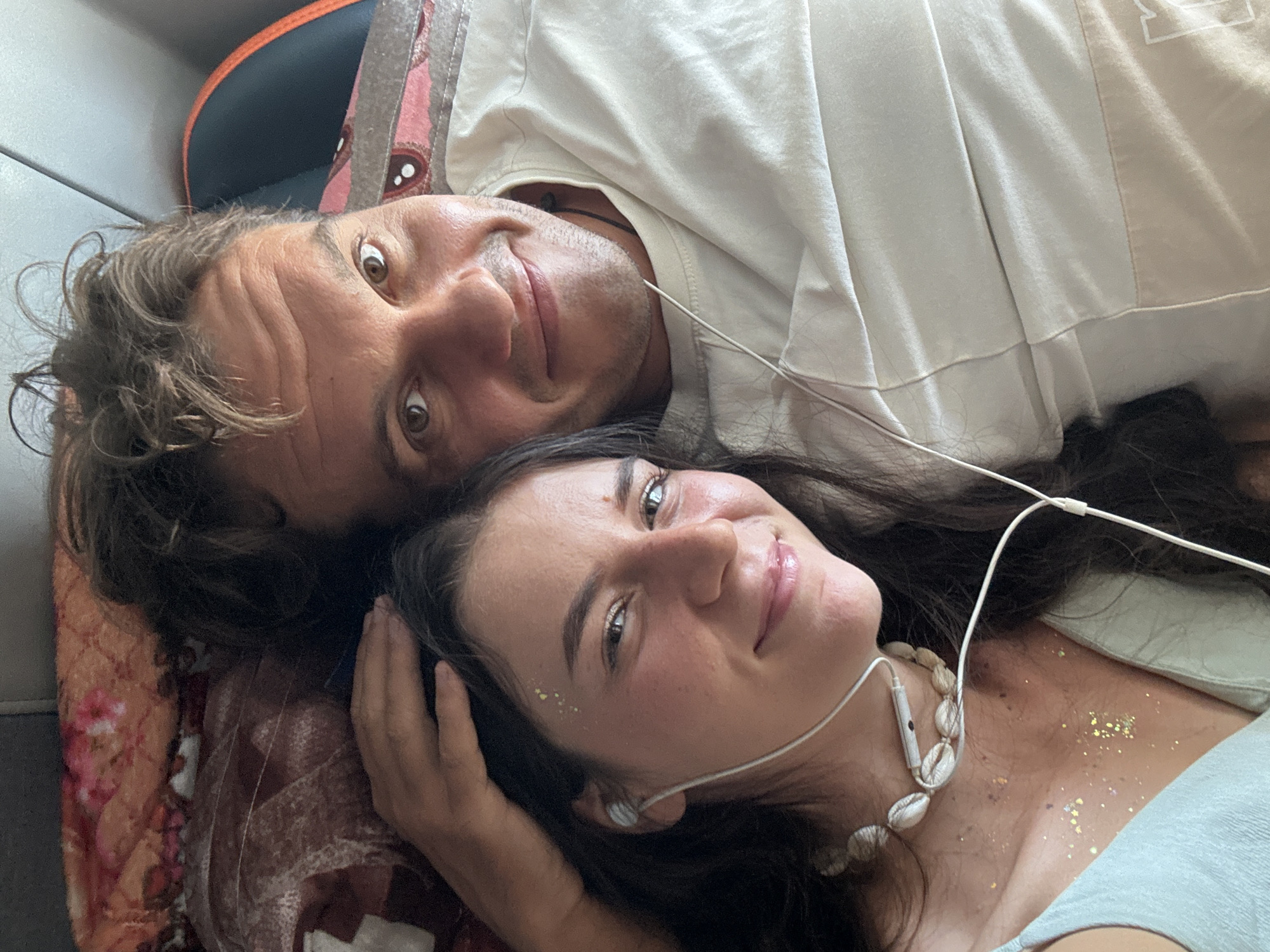 Two individuals, a man and a woman, are lying down, possibly indoors. The man is positioned above and to the left of the woman. Both appear to be looking at the camera with slight smiles. They have earphones in their ears. The woman wears a shell necklace and has glitter on her neck and collarbone. They appear to be on a patterned fabric or bed covering. The photograph may have been taken in Phu Thuy Ward, Vietnam.