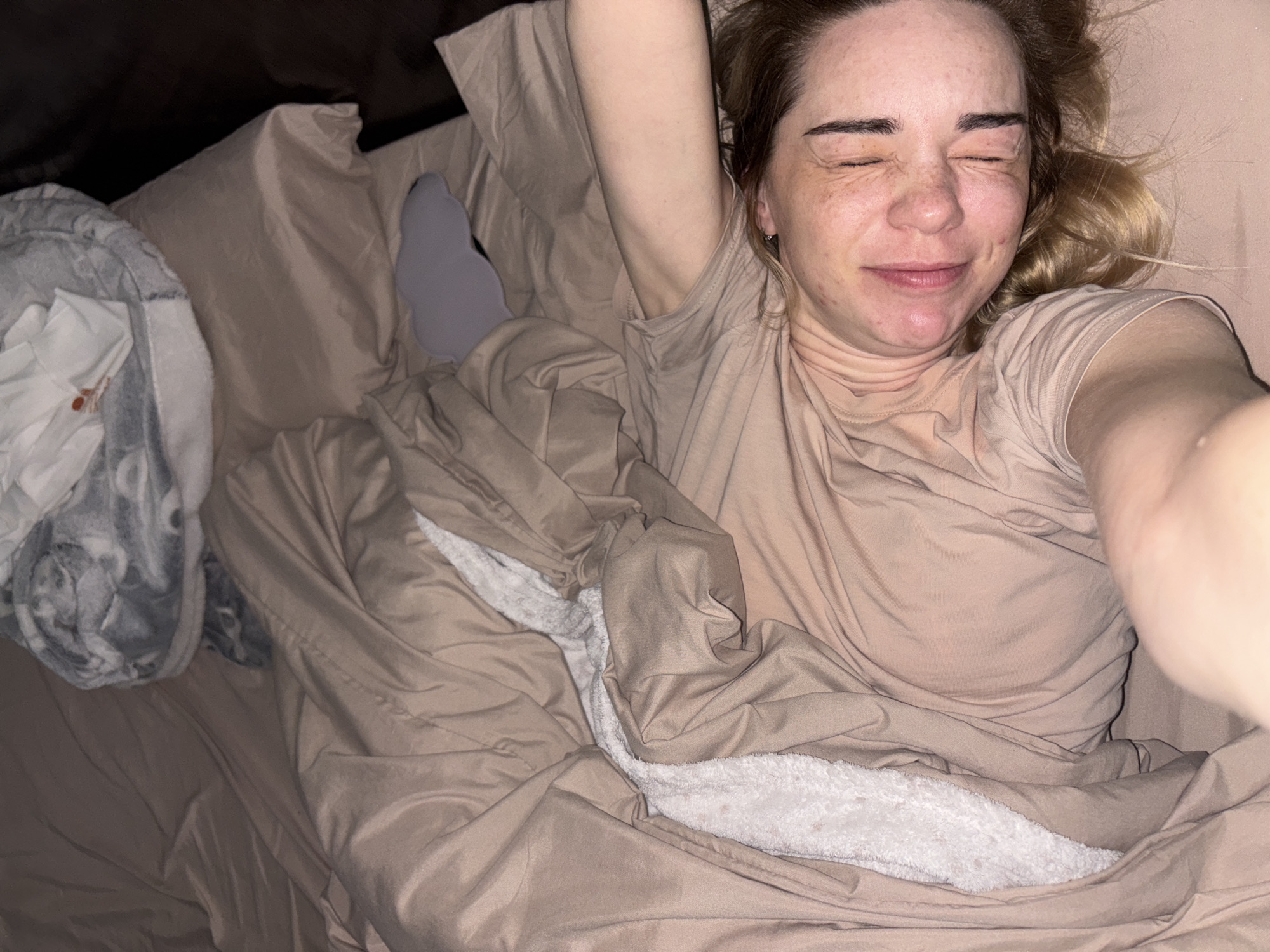 A woman with freckles is lying in bed. She has blonde hair and is wearing a tan t-shirt. She is smiling and her eyes are closed. She is covered in a tan duvet with a white plush lining. There is a grey pillow on the left side of the image. The location is unknown, Russia.