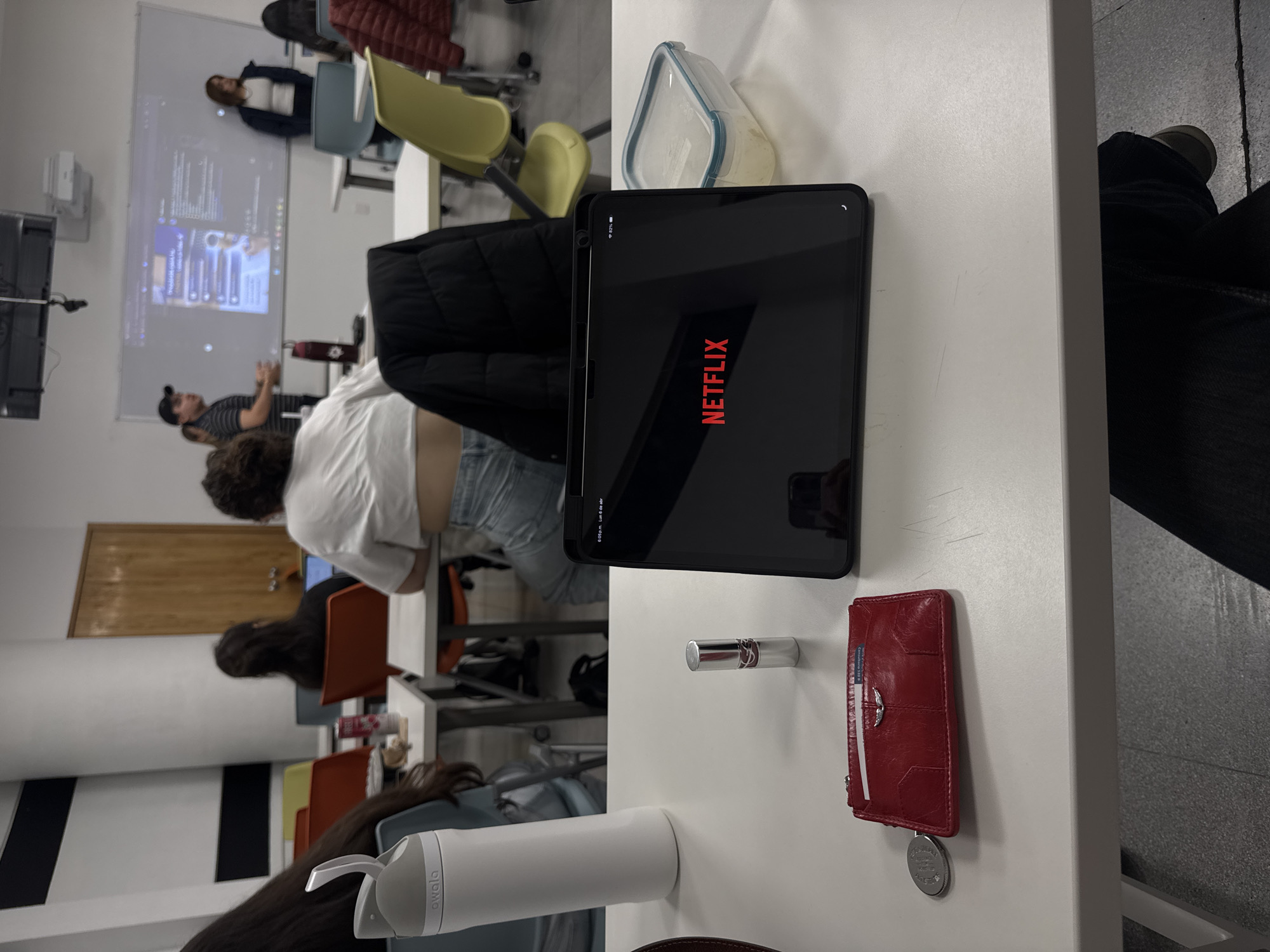 The image depicts a classroom scene, possibly a university in Puebla, Mexico. People are seated at desks facing a whiteboard displaying a presentation. A student in the foreground is watching Netflix on a tablet during class. Other objects on the desk include a water bottle, a food container, a lipstick, and a red wallet. A jacket is draped over the back of a chair. The floor is tiled.
