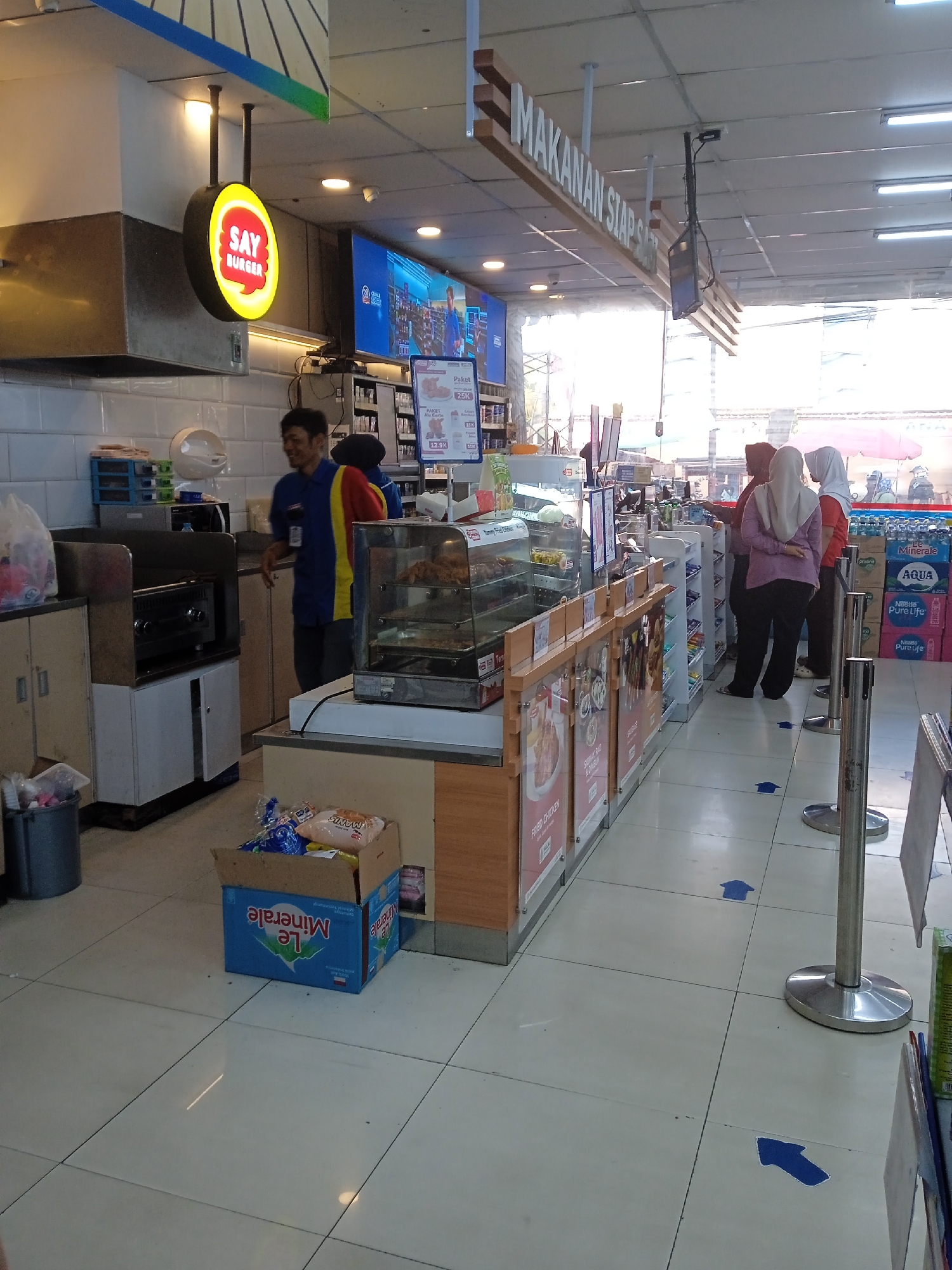 The image depicts the interior of a convenience store in Kaliwungu, Indonesia. A circular sign with yellow and red lighting, bearing the text "SAY BURGER," is visible above a food counter. A store employee, wearing a blue and yellow uniform, stands behind the counter, smiling and looking to the left. To the right of the employee, a glass display case holds fried chicken. Further down the aisle, several customers are queuing, indicated by blue arrow floor markings. One customer is wearing a light purple hijab and a dark top, another a white hijab and an orange top, and a third is partially obscured. Shelves stocked with various products, including bottles of "Aqua Pure Life" and "Le Minerale," line the right side of the store. The store has a tiled floor and a suspended ceiling with fluorescent lighting. The overall scene suggests a typical retail environment with customers engaged in shopping and staff providing service.