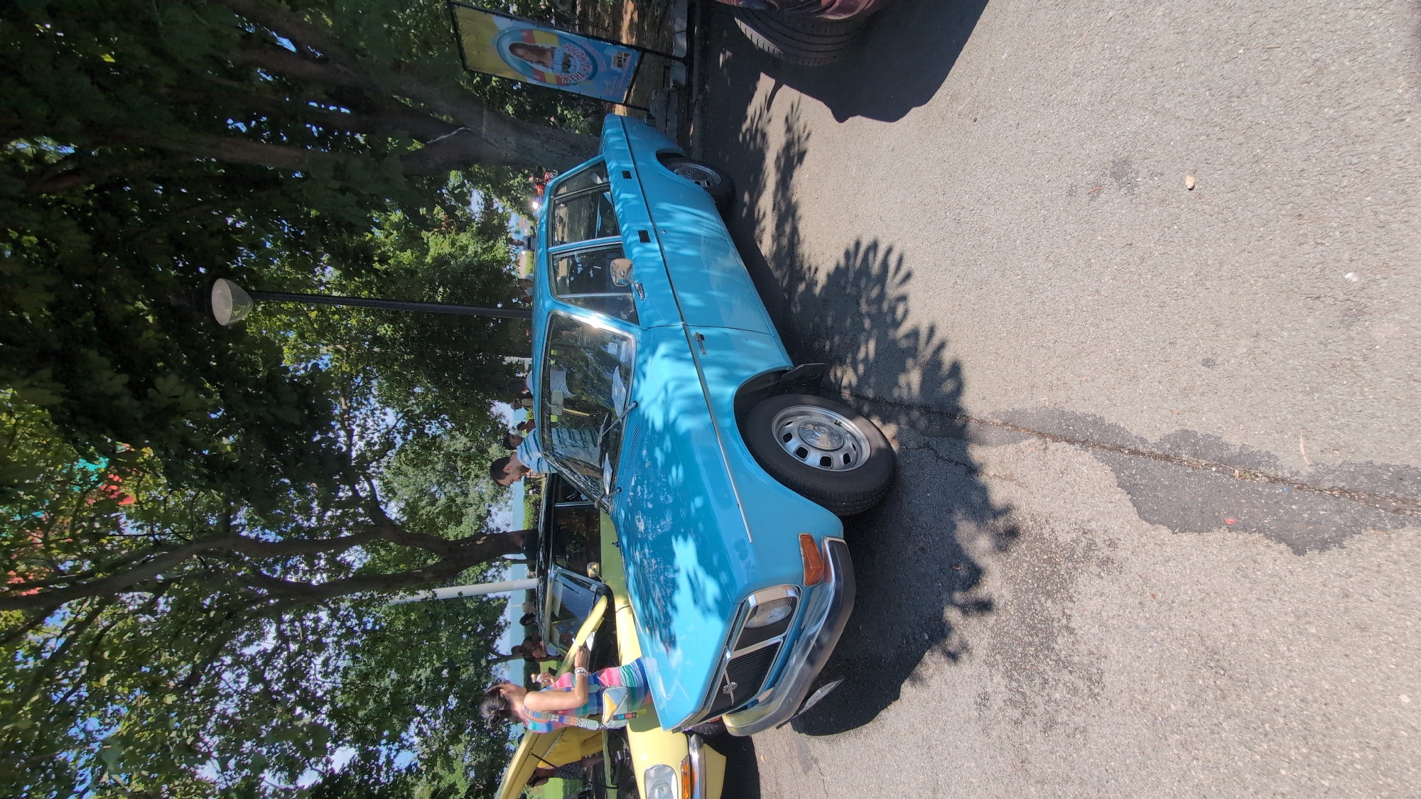 An eye-level, full shot captures a vintage light blue Volvo sedan parked on an asphalt surface, likely during a car show or outdoor event. The car is angled, with its front-left quarter most prominent. The vehicle's chrome bumper and grille are visible, as are its silver hubcaps and the side profile. Behind the Volvo, a yellow car with its hood up is partially visible, with a woman in a colorful dress standing near it. In the background, several people are gathered, with trees providing ample shade. A lamppost stands behind the Volvo. To the right, a colorful poster with a woman's image is propped up. The scene suggests a warm, sunny day. The location is identified as the Sea Garden in Burgas, Bulgaria, indicated by the signage of the "Sea Garden" and the characteristic coastal trees.