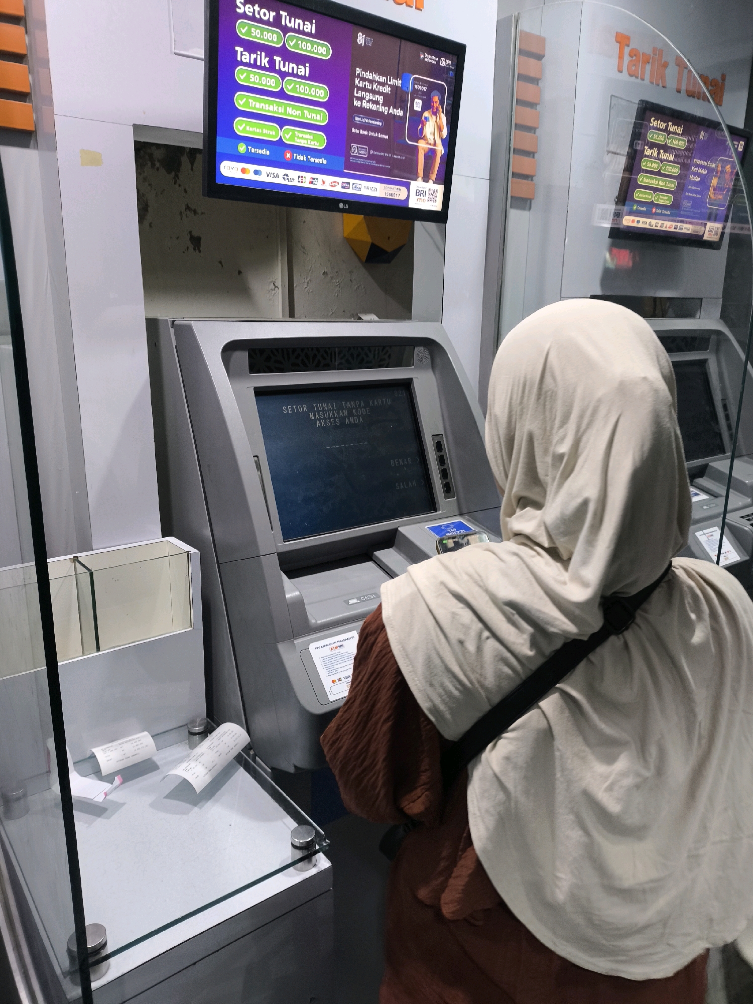 The image depicts a person from behind, wearing a light beige hijab and a brown tunic, standing in front of an ATM in Limboto, Indonesia. The ATM screen displays options for cash deposit and withdrawal, with available denominations of 50,000 and 100,000 Indonesian Rupiah. Below these options, there are icons for various card networks like Visa, Mastercard, and others. To the left of the ATM, there is a glass enclosure containing several printed receipts. Another ATM screen is visible to the right, also displaying "Tarik Tunai" (Cash Withdrawal) and transaction options. The overall environment suggests a banking or financial service location.