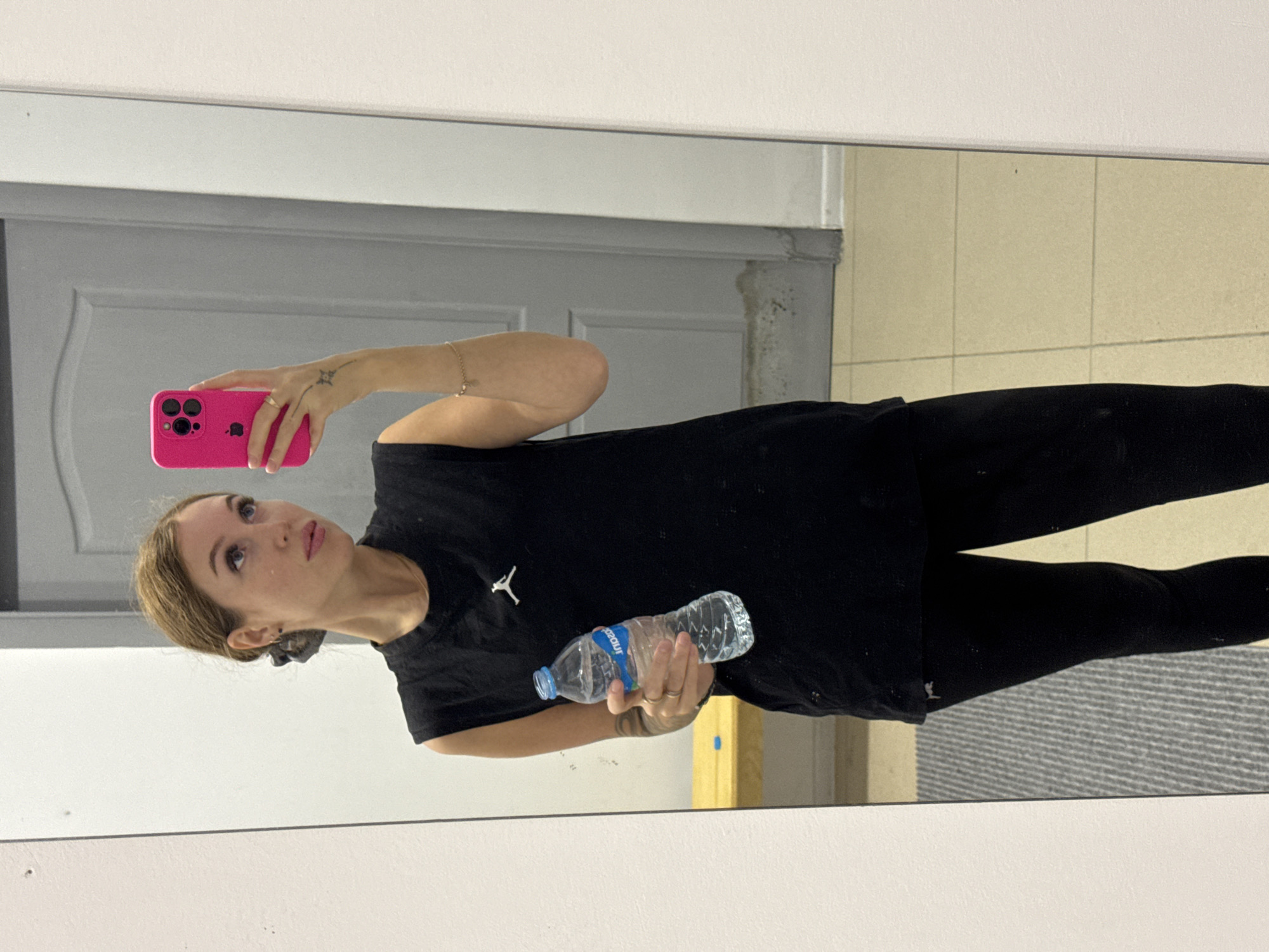 A young woman is captured in a mirror selfie, taken indoors in Chaikovsky, Russia. She is shown from the waist up, looking towards the pink smartphone she holds up in her right hand to take the picture. In her left hand, she holds a clear plastic water bottle with a blue label that appears to read "AQUAZOU".
She is wearing a black short-sleeved t-shirt with a white Jordan Jumpman logo on the left side of the chest, and black leggings. Her light brown hair is pulled back, possibly with a dark scrunchie, and she has a neutral expression, gazing at the phone's screen. A ring is visible on her right ring finger, and a delicate bracelet adorns her left wrist. A small, subtle tattoo is visible on her right forearm.
The reflection in the mirror reveals a straightforward indoor environment. Behind the woman, a light-colored wall rises, and to her left, a grey door with molded paneling is visible. To her right, the reflection shows a beige tiled floor and a grey textured mat or rug. The lighting suggests a well-lit interior, implying the photograph was taken during daylight hours or in a brightly illuminated room. The scene depicts a casual moment, possibly within a gym, changing room, or at home.