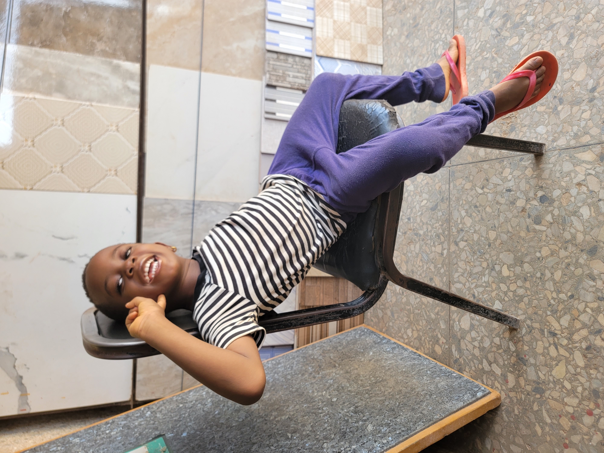 A young child lies upside down on a black chair, laughing and looking toward the camera. The child wears a black and white striped shirt, purple sweatpants, and coral-colored flip-flops. The chair is positioned against a wall covered in tile samples of various colors and patterns. The floor is tiled with a speckled pattern. A gray table with a wooden edge is visible under the chair. The image appears to be taken in a retail environment, potentially a tile store in Maiburiji, Nigeria.