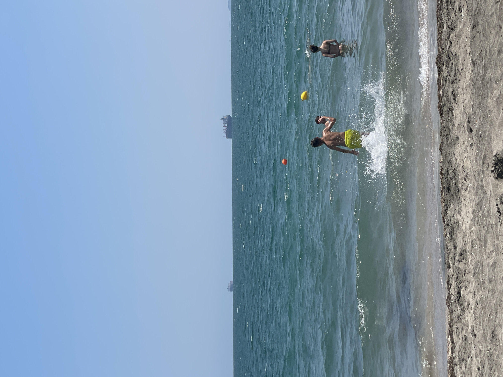 The image depicts a sunny day at the Black Sea beach in Burgas, Bulgaria. In the foreground, wet sand with some debris lines the shore. The sea is a vibrant turquoise, with gentle waves breaking on the beach. In the water, two young people are visible. One, wearing yellow swim shorts, is facing away from the camera and appears to be playing in the water, with a splash of water behind them. Another person, wearing a dark bikini, is standing further out in the water. Two buoys, one orange and one yellow, are floating in the sea. In the distance, several large cargo ships are visible on the horizon, under a clear blue sky.