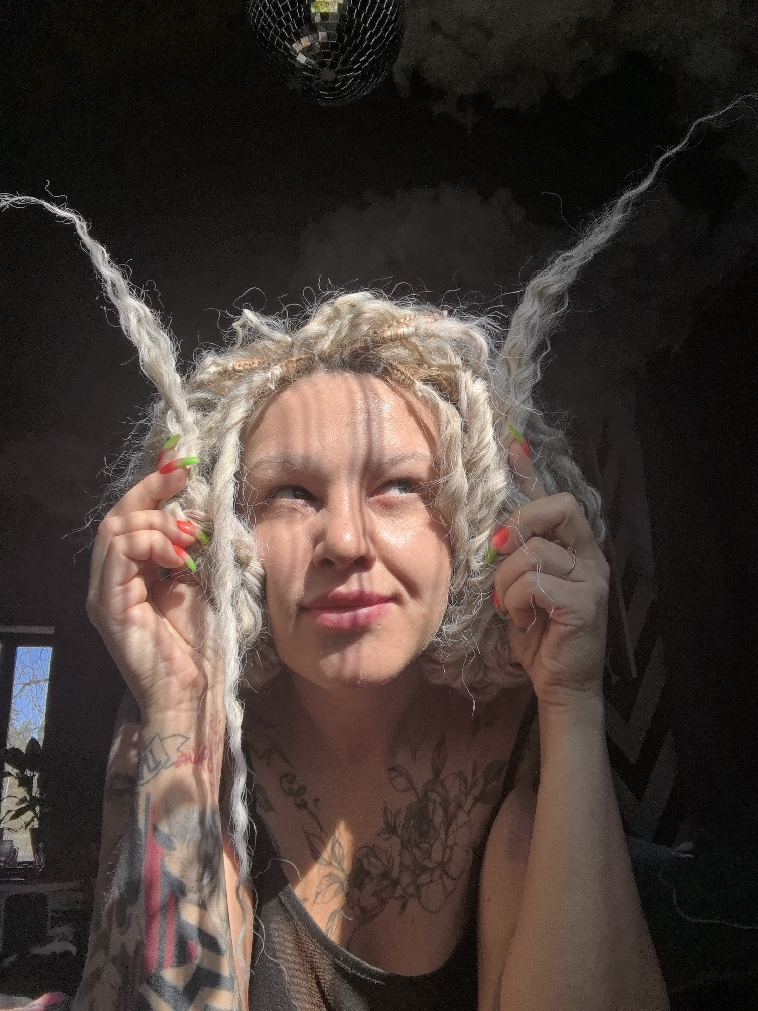 A woman with blonde and light-colored dreadlocks is the central subject of this image, captured in an indoor setting in Noviy, Russia. She looks upward with a subtle, contemplative expression, holding strands of her unique hair near her temples with both hands. Her long, pointed nails are distinctively painted with a vibrant red-to-green gradient. Tattoos are visible on her arms and chest, including a prominent floral design on her left shoulder/décolletage. She is wearing a dark tank top.
The scene is illuminated by strong, direct sunlight streaming in from an unseen source, creating distinct striped shadows across her face and upper body, highlighting certain features while leaving other areas in shadow. In the upper background, a disco ball is partially visible, reflecting specks of light. To the left, a window provides a glimpse of the outside world, showing bare tree branches and blue sky, with a houseplant visible indoors near the sill. The background of the room is mostly dark, with some faint, geometric patterns (possibly chevron or striped) visible on a wall to the right. The strong sunlight suggests it is daytime. No visible text is present in the image.