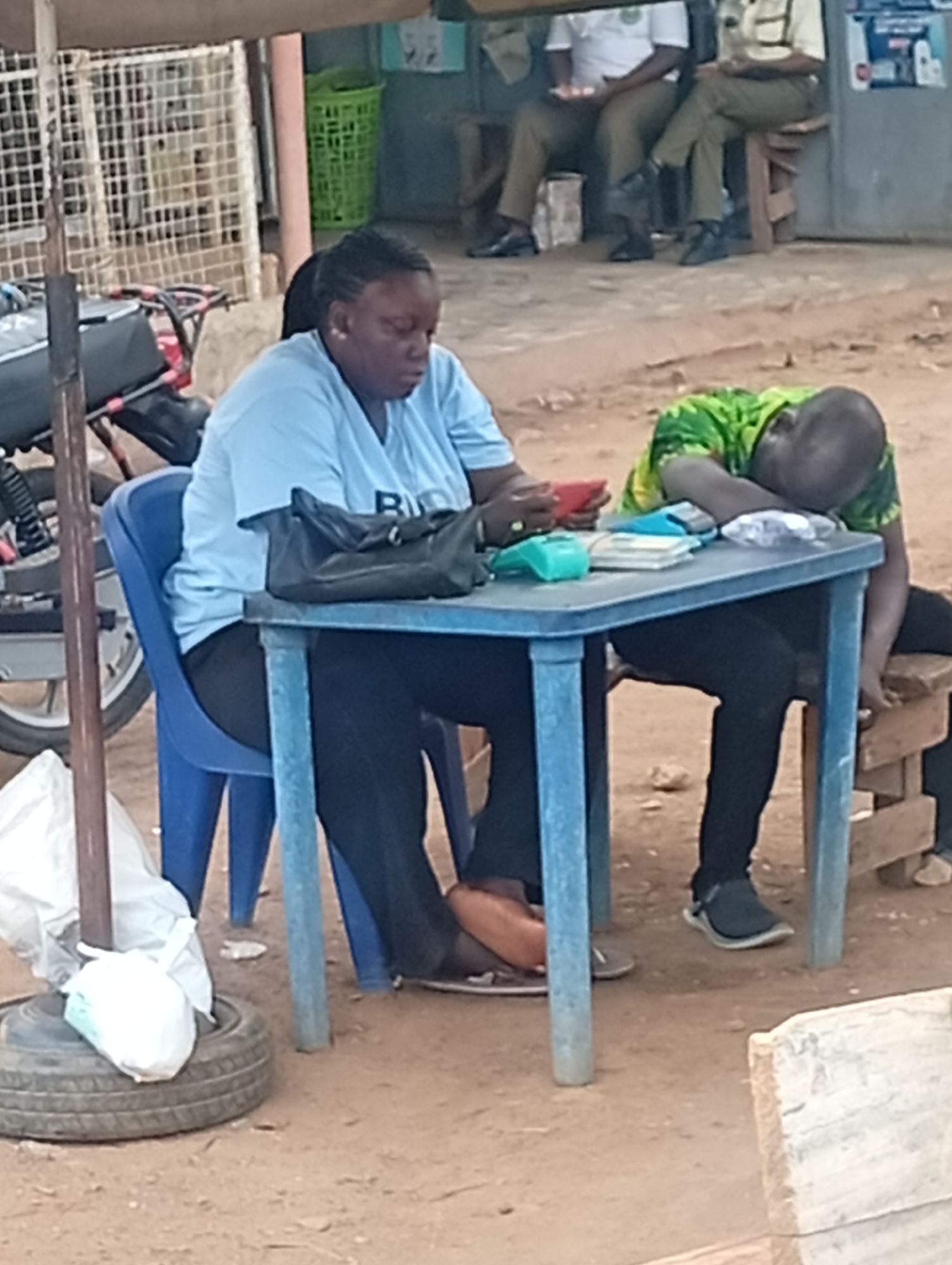 In Sabon Tasha, Nigeria, a woman in a light blue shirt and dark pants is seated at a light blue plastic table outdoors, looking at a red mobile phone in her hands. On the table is a black purse, a green payment device, and a stack of cash. Seated at the same table is a person wearing a green and yellow shirt, resting their head on the table. Behind the woman is a blue plastic chair and a parked motorcycle. In the background, two men are seated on a bench in front of a building. A tire is on the ground in the foreground, supporting what appears to be a pole.