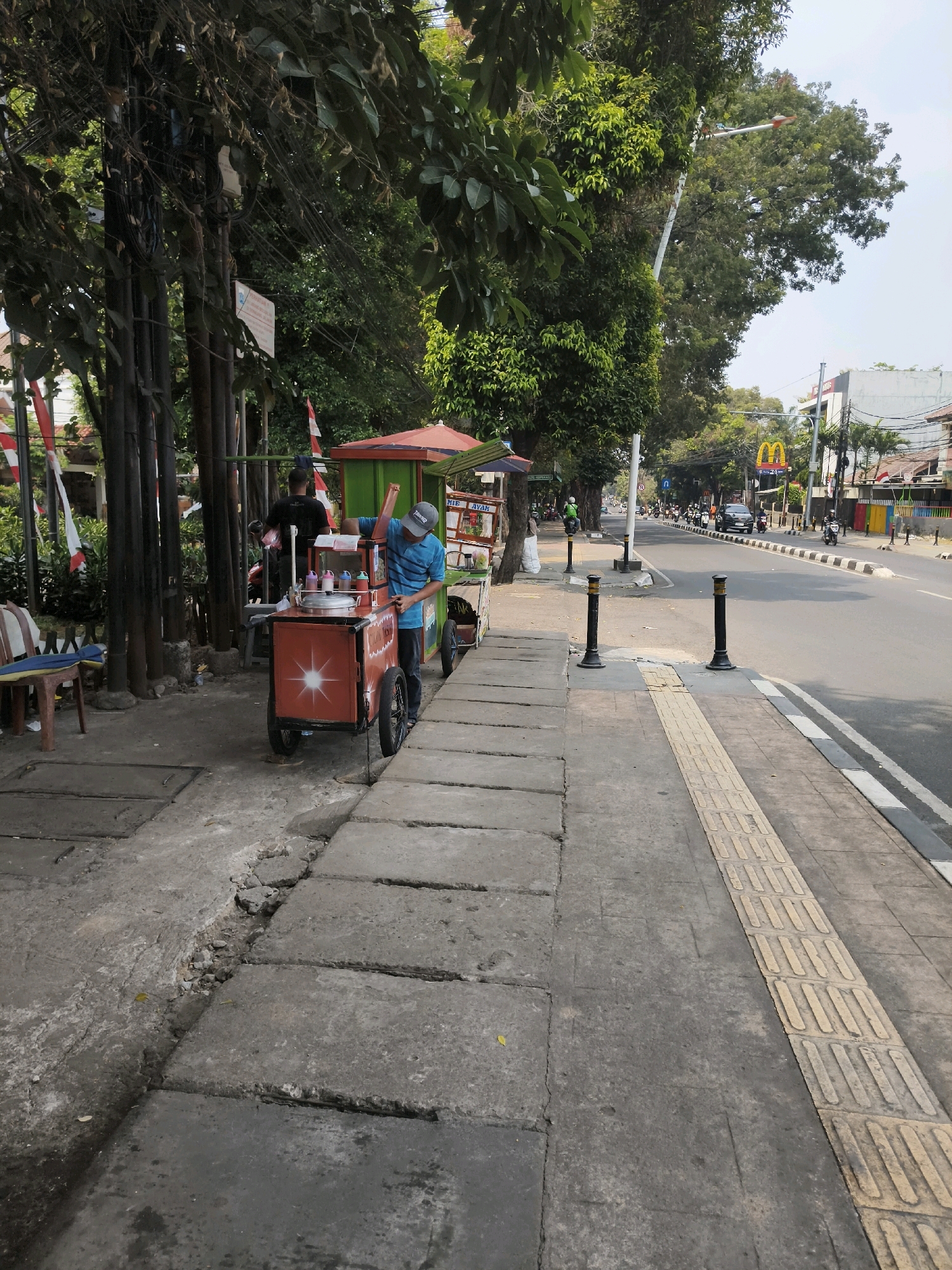 This image depicts a street scene in South Jakarta, Indonesia. On the left, a sidewalk is lined with trees, electrical poles, and a food cart vendor. The vendor, a man wearing a blue striped shirt and a cap, is interacting with his cart, which is orange and red with a white star design and wheels. The cart appears to be selling a cold dessert, with various colored liquids in containers on top. Behind him, another person is standing near a darker colored cart. There are also chairs and tables on the sidewalk. Red and white flags are visible in the background. The sidewalk transitions to a textured, yellow tactile paving for the visually impaired, which runs alongside the street. The street itself is asphalt, with white and black lane markings. Traffic, including motorcycles and cars, is visible on the street. A McDonald's sign is visible in the distance on the right side of the road. The environment appears to be a commercial or residential area with buildings and more trees lining the street. The weather is sunny with a clear sky. The condition of the sidewalk shows some signs of wear and tear, with cracked concrete slabs.