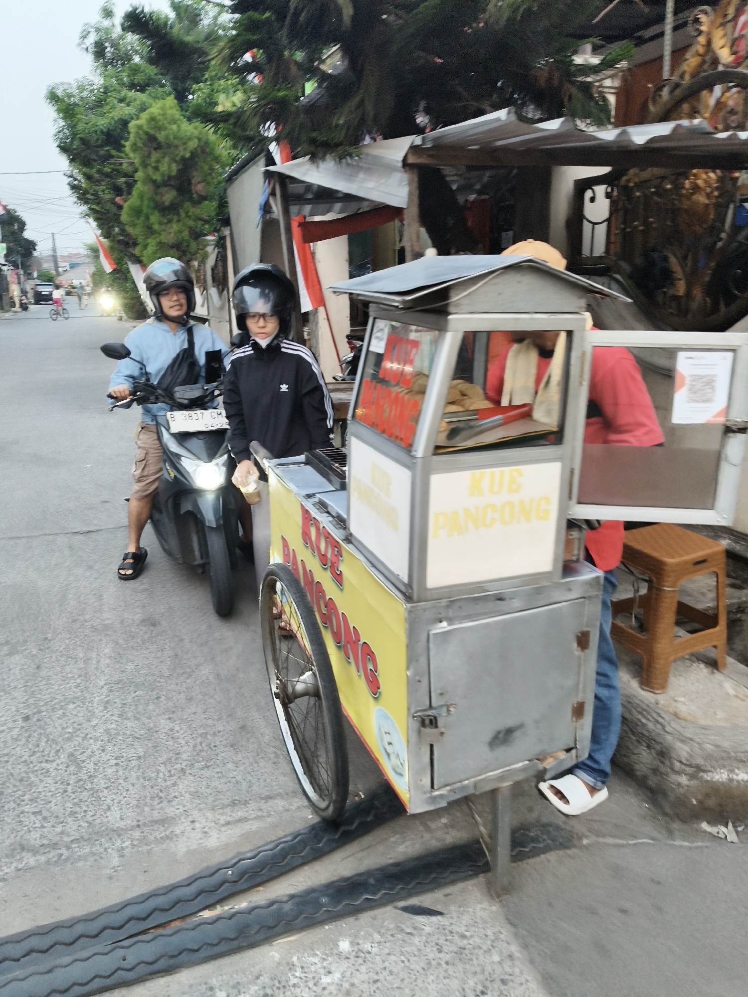 In South Tangerang, Indonesia, a person wearing a light blue long-sleeved shirt and beige cargo shorts, identified as male, rides a black scooter with a license plate reading "B 3837 CM". He is wearing a black helmet. A person wearing a black jacket with white stripes on the sleeves and a white face mask, identified as likely female or young male due to facial features and build, sits behind him on the scooter, also wearing a black helmet. They are stopped next to a yellow and silver food cart labeled "KUE PANCONG" in red and yellow lettering. The food cart has a glass display case showing food items, cooking utensils, and a QR code displayed on a white panel. A person wearing a red t-shirt and blue jeans is standing behind the food cart, partially visible. The environment is a paved street with some unevenness in the asphalt, including some raised black rubber speed bumps. Trees and buildings are visible in the background, with Indonesian flags decorating one of the structures. The sky is overcast.