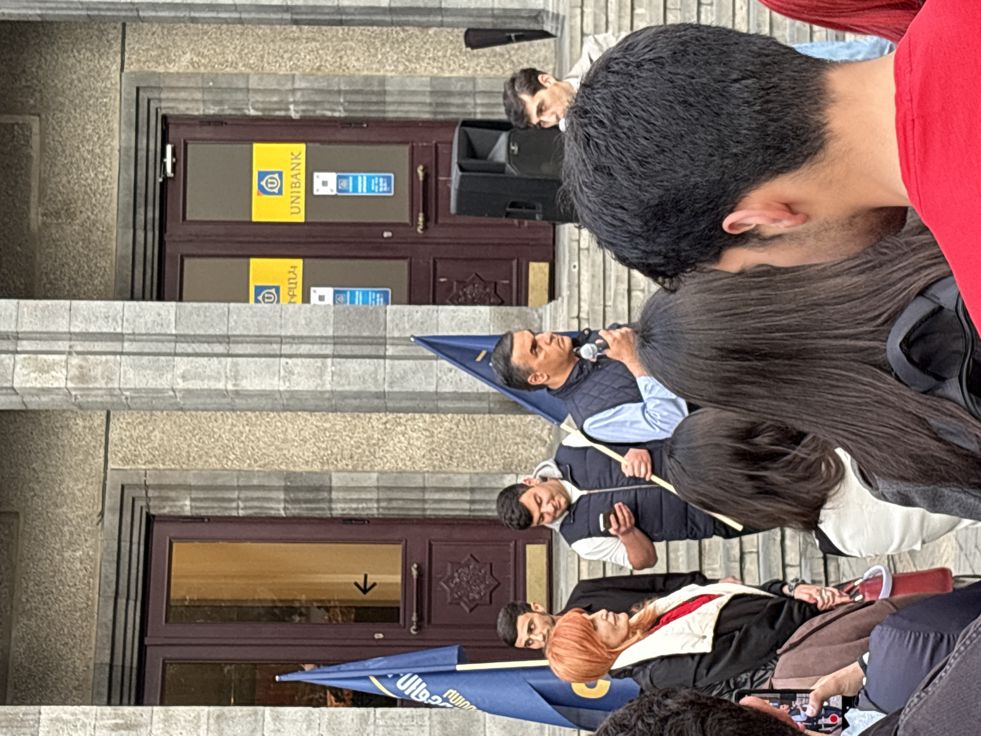The image captures a scene in Yerevan, Armenia, in front of a building with "UNIBANK" signage on its doors. A group of people are gathered, with at least one man speaking into a microphone. Some individuals hold blue and yellow flags. The architecture appears to be Soviet-era. The focus is primarily on the people and their actions within the urban environment.