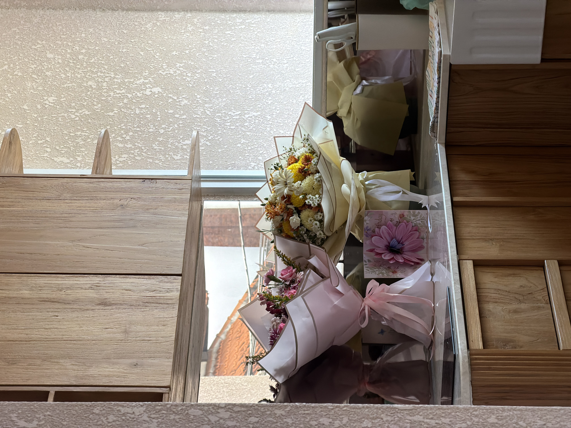 The image shows a view from what appears to be an elevated indoor location, potentially a window or a balcony, overlooking a cityscape in Mengwi, Indonesia. Two bouquets of flowers, one with white and yellow blooms wrapped in beige paper and the other with pink and purple flowers wrapped in pink paper, are positioned in front of the window or balcony. A birthday card with a pink flower is also visible. The external view includes rooftops with red tiles, suggesting a residential area. The interior space seems to be furnished with wooden elements, including what looks like a set of drawers.