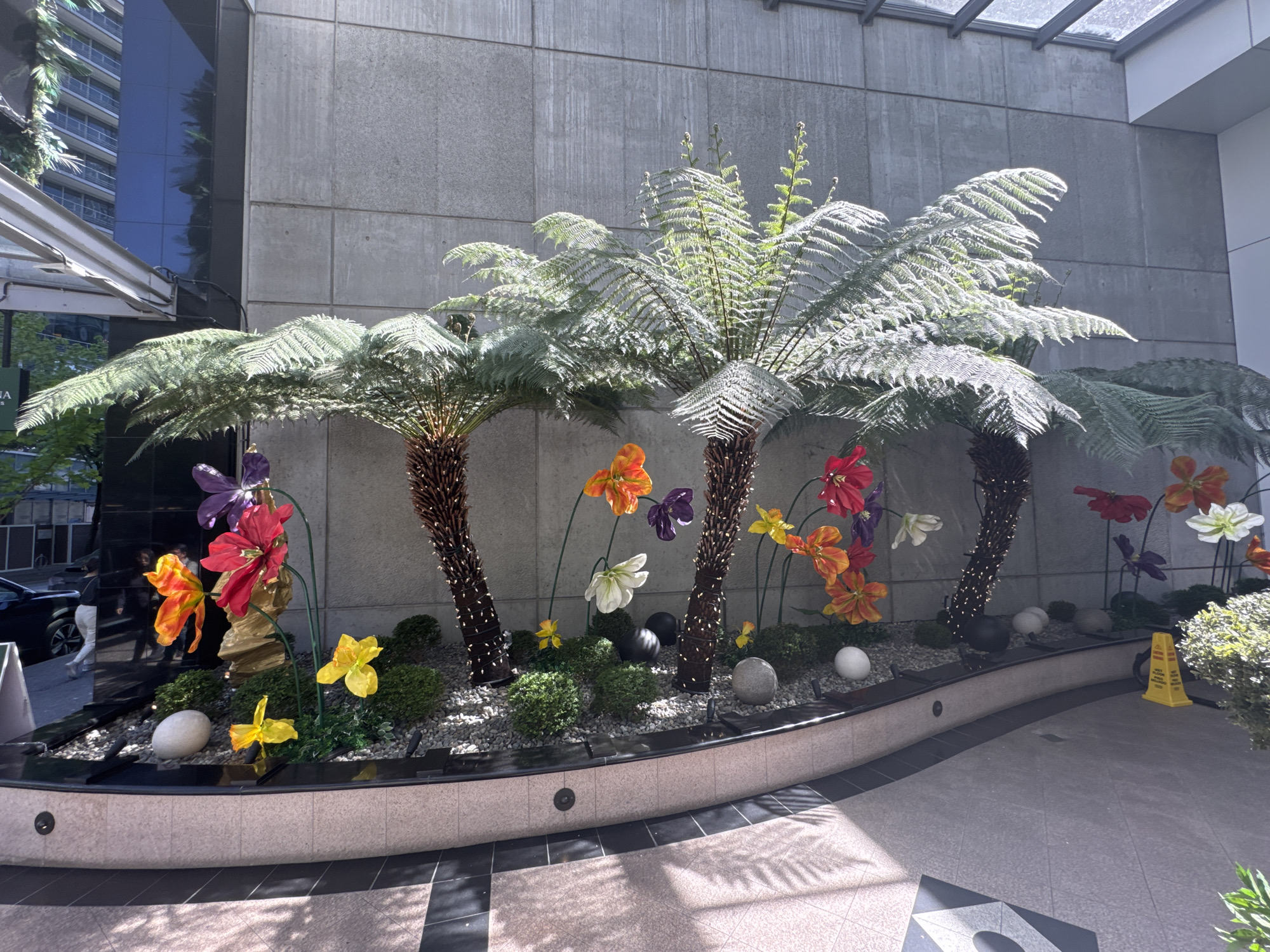 The image depicts a decorative arrangement of artificial tree ferns and colorful artificial flowers within a curved planter, situated in what appears to be a courtyard or entrance area in Vancouver, Canada. Three tree ferns with their distinctive fronds dominate the scene, each wrapped with what appear to be strings of small lights. Scattered amongst the ferns are colorful, large-scale artificial flowers in shades of orange, red, purple, yellow, and white. The planter bed is filled with small pebbles, green bushes, and scattered decorative spheres in shades of white, gray, and black. The planter itself is finished with a stone-like surface. The background features a concrete building facade and a glimpse of a taller building with reflective windows. Parts of the floor are covered in geometric black and gray patterned tile and there is a yellow traffic cone on the right side of the image. A few figures can be seen out of focus, behind the foliage on the left.