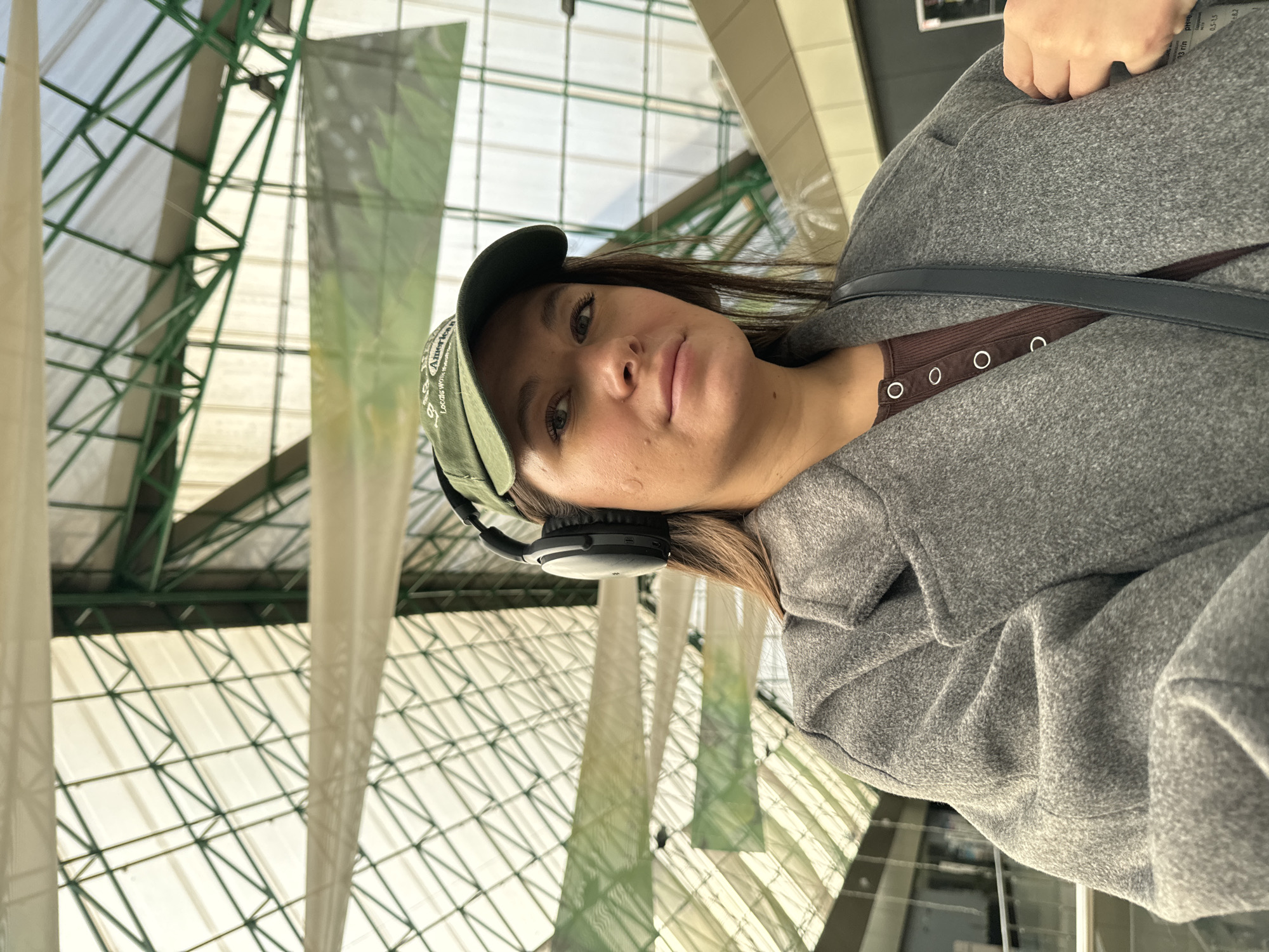 A female individual is depicted in a medium shot, centered slightly to the right, looking directly at the camera with a neutral expression. She wears a dark olive green baseball cap with partially visible white embroidered text "Locals work American" and black over-ear headphones. Her attire includes a grey textured long coat worn over a dark brown ribbed top with three visible silver-ringed buttons. A dark strap, consistent with a shoulder bag, crosses her left shoulder. The background features a large indoor space characterized by a geometric ceiling structure composed of green metal beams and translucent panels, allowing ambient light. Several light-colored rectangular banners hang horizontally in the mid-ground. To the right, a blurry white sign or display board with indistinct text and numbers (e.g., "3 r/m", "pH", "0.5-1.5") is partially visible. This scene is located within a public facility in the city of Odintsovo, Russia.
