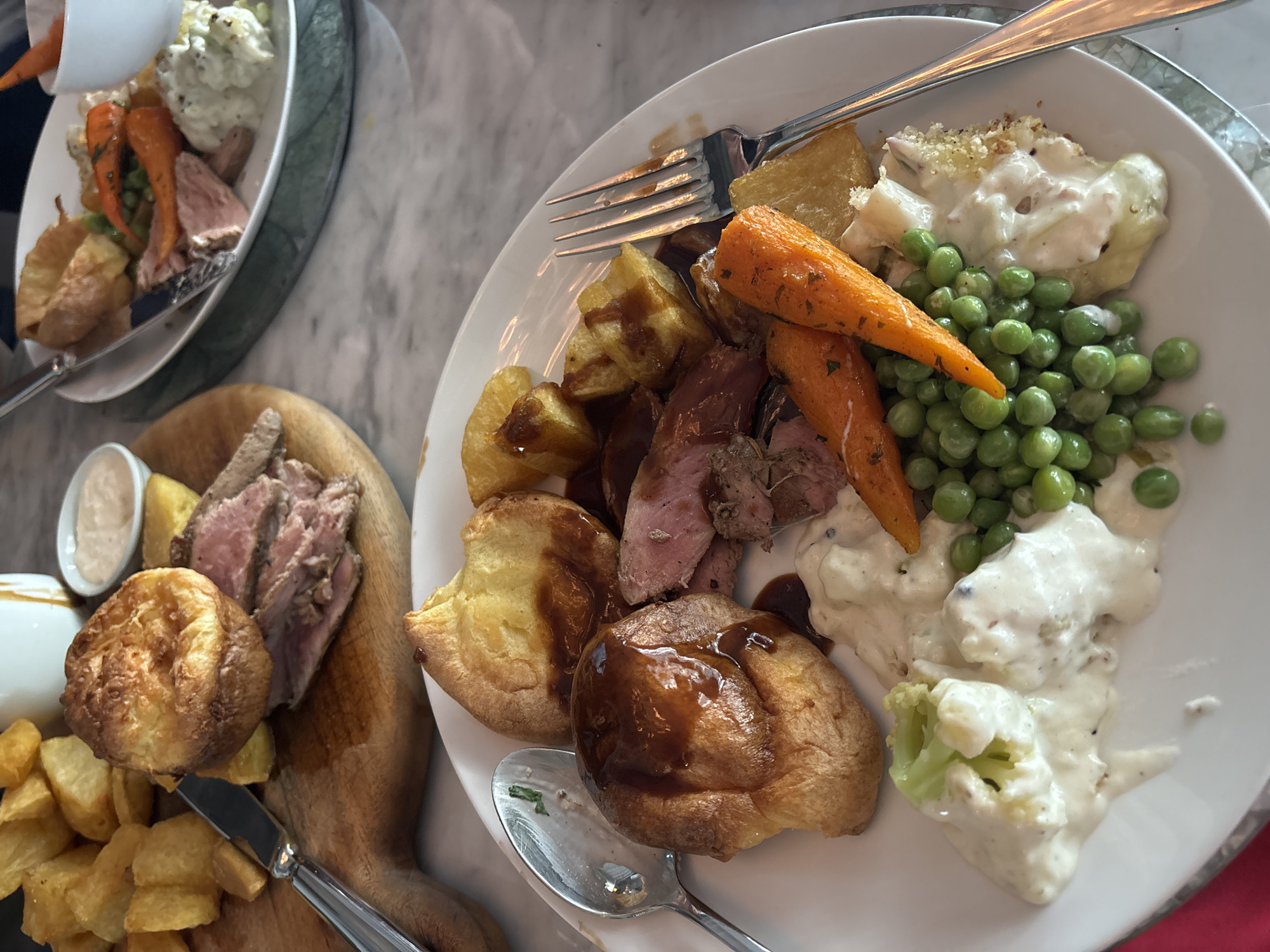 A high-angle, close-up shot showcases a table laden with food. On the right, a white plate is piled with roast beef, Yorkshire pudding topped with gravy, roasted potatoes, carrots, peas, and cauliflower cheese. A fork and spoon lie atop the plate. To the left, a wooden board displays sliced roast beef, a Yorkshire pudding, and what appear to be chips, alongside a small white dish filled with sauce. Above, another white plate contains a similar meal, including meat and vegetables. The background reveals a light, marble-like surface. The setting is indoors, and the presence of cutlery suggests a meal in progress. The city of North Kuta, Indonesia, is not visibly relevant in this image.