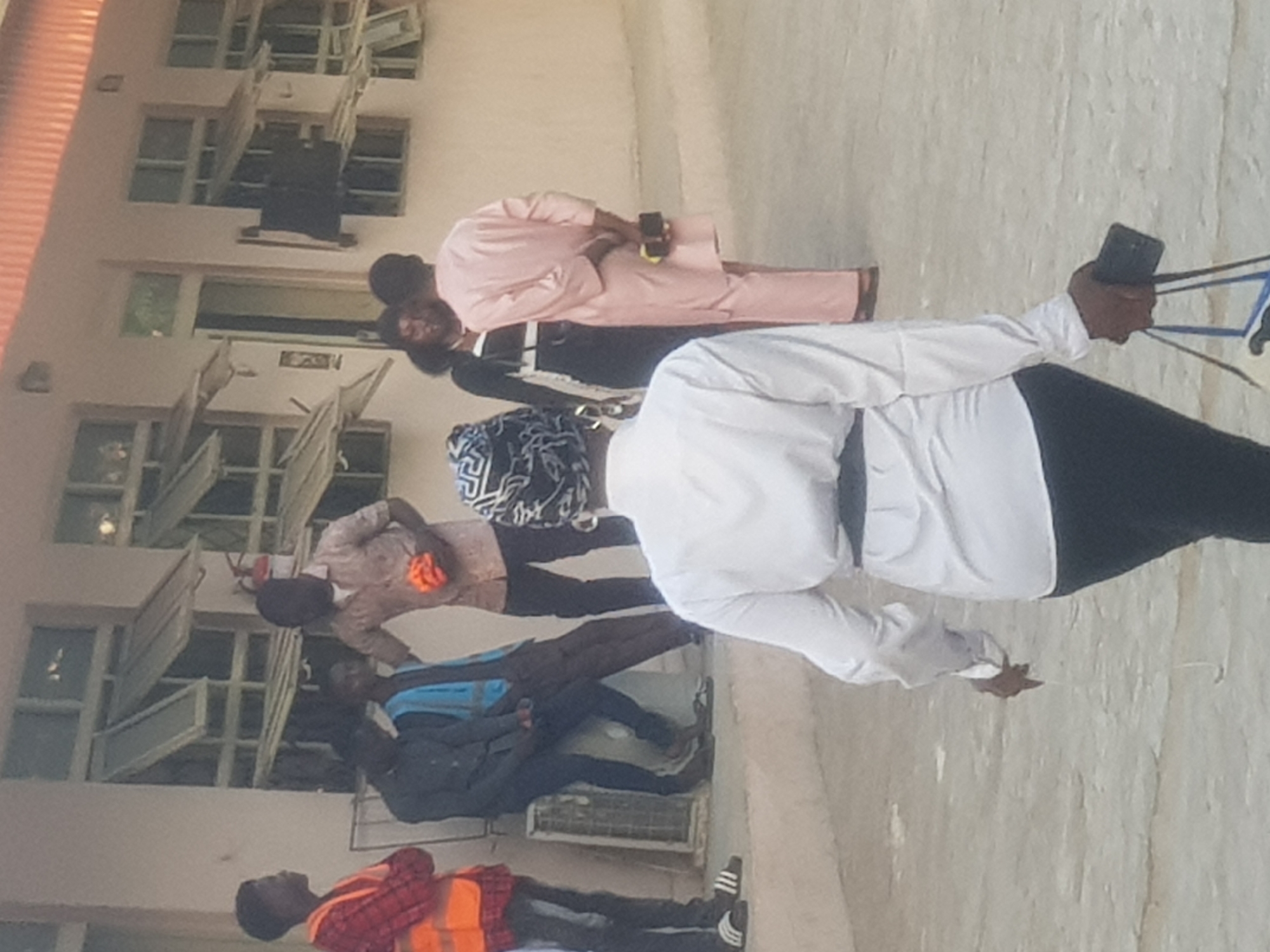 The image depicts a group of people standing in front of a building in Dabawa, Nigeria. The building has multiple open windows. The group is diverse in attire; one individual wears a pink suit, another a white shirt and black pants, and a third a reflective safety vest. Some members of the group appear to be holding phones. There is debris on the ground.