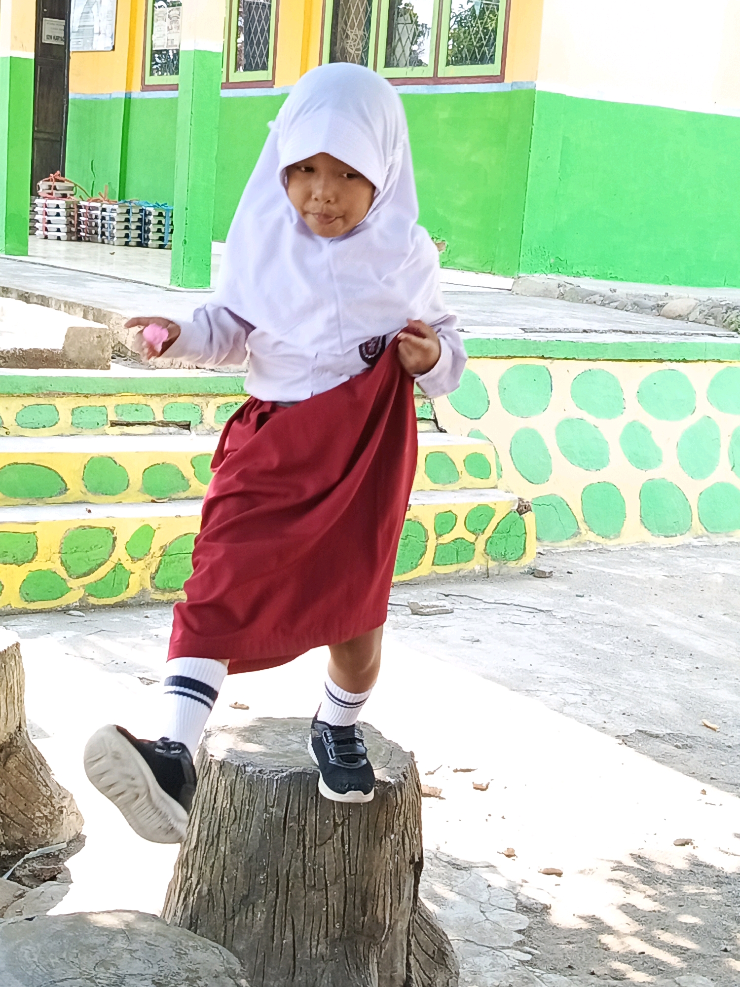 A young girl, wearing a white hijab and a school uniform consisting of a white shirt and a red skirt, stands on a weathered tree stump. Her white socks have dark stripes. She holds a small pink object in her left hand. Her right foot is raised, and her right leg is bent at the knee. She is facing forward, looking slightly to her right.
The background features a school building painted in yellow and bright green, with green window frames. There are steps leading up to the building, painted with yellow and green polka dots. In the distance, stacks of white and red trays are visible. The ground is paved with concrete, with some scattered dry leaves. The scene is outdoors, with sunlight casting shadows on the ground. The location is identified as Karya Dadi, Indonesia.