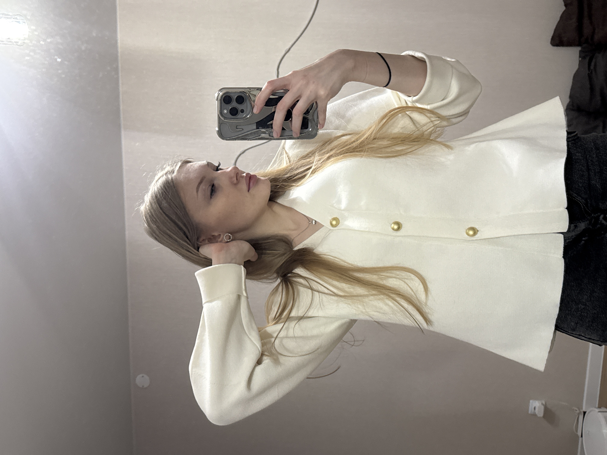 A woman stands in a room, possibly in the city of Perm, Russia, holding a smartphone to take a selfie in a mirror. She wears a white cardigan with gold buttons and dark-colored pants. Her blonde hair falls over her shoulder. The environment includes a light-colored wall and possibly a wardrobe or hanging clothes visible in the background.