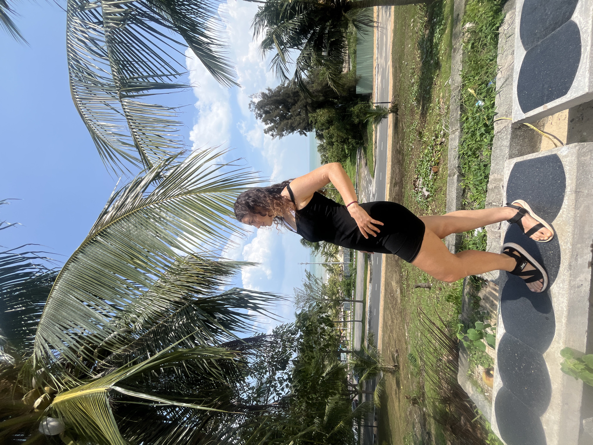 A full-shot photograph captures a woman standing on a concrete ledge, facing right, looking out at the ocean. She is wearing a black sleeveless dress and black and white sandals. Her curly brown hair is worn down. The woman is standing in front of a backdrop of palm trees, a street, and a beach. The sky is blue with scattered clouds. The scene is likely in a coastal city in Vietnam, given the presence of palm trees and the ocean view.