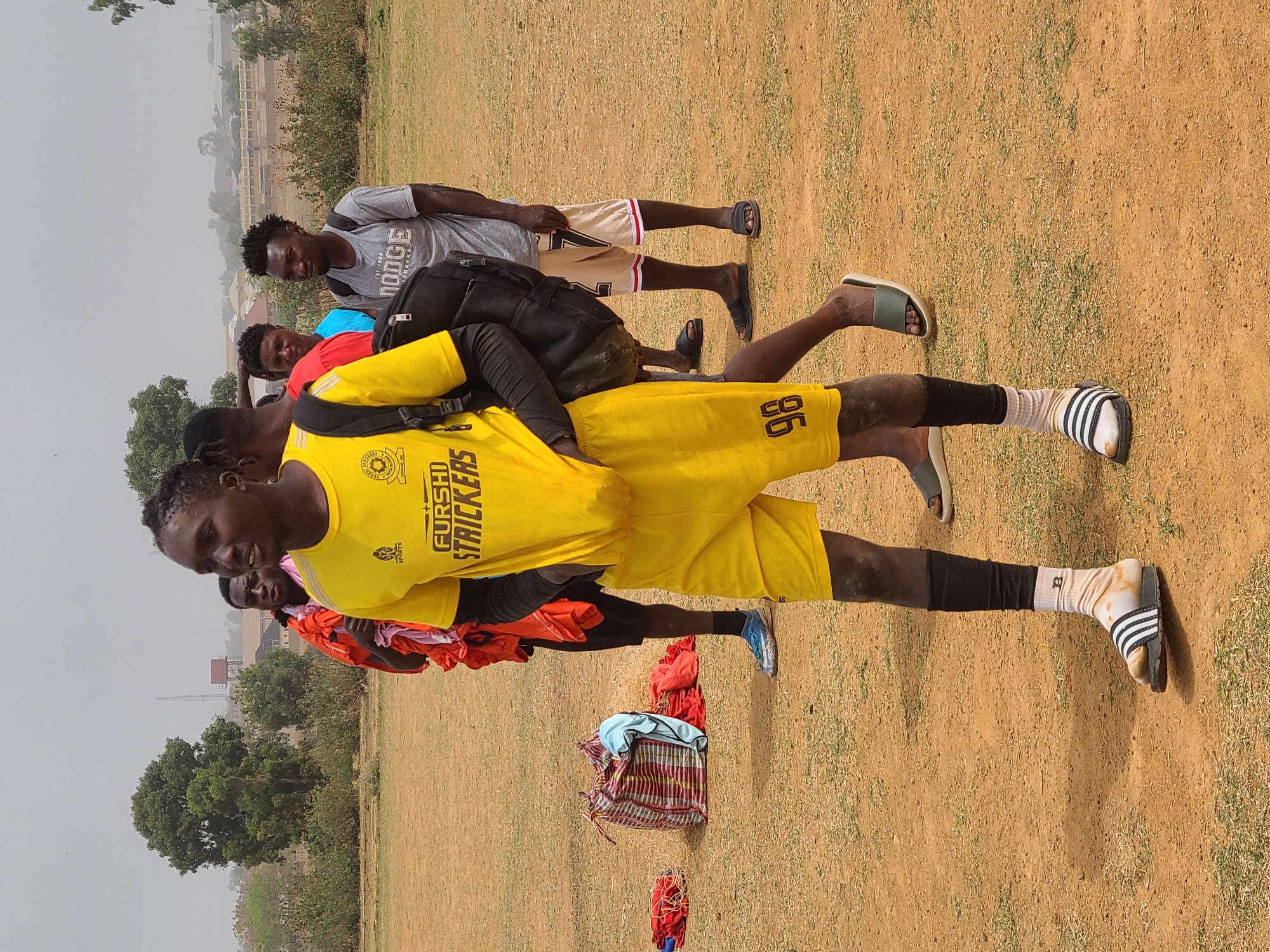 This portrait-oriented outdoor photograph depicts a group of young men on a dusty, unpaved field with sparse patches of dry grass, under a hazy sky. The setting appears to be a training ground or a casual sports area in Barikin Jirga Summa, Nigeria.
In the foreground, a prominent male figure with dark complexion is captured from the waist up. He is wearing a bright yellow soccer jersey and matching shorts. The jersey has "FURSUN STRICKERS" printed vertically on the right side and a "SPORTS" logo on the left chest. The number "96" is visible on the back of his shorts. He also wears black compression sleeves on his calves, white socks with black stripes, and black and white striped slides (Adidas brand). A black backpack is slung over his shoulders. His expression is neutral as he looks slightly downward.
Behind him, several other men are visible. One man wears a grey t-shirt with "DODGE" and "EST. 1888 NEBRASKA" printed on it, paired with khaki shorts and black slides. Another individual in the mid-ground wears a red shirt, and another a blue shirt. The men appear to be gathered, suggesting they are resting or preparing for an activity like a soccer game or training session.
On the ground near the main subject, there is a pile of clothes or fabric, including a red and white striped bag, a blue item, and a red mesh bag, possibly containing sports equipment or personal belongings. In the background, scattered trees and distant, indistinct buildings or structures are visible under the same hazy conditions, reinforcing the outdoor, potentially rural or semi-urban environment.