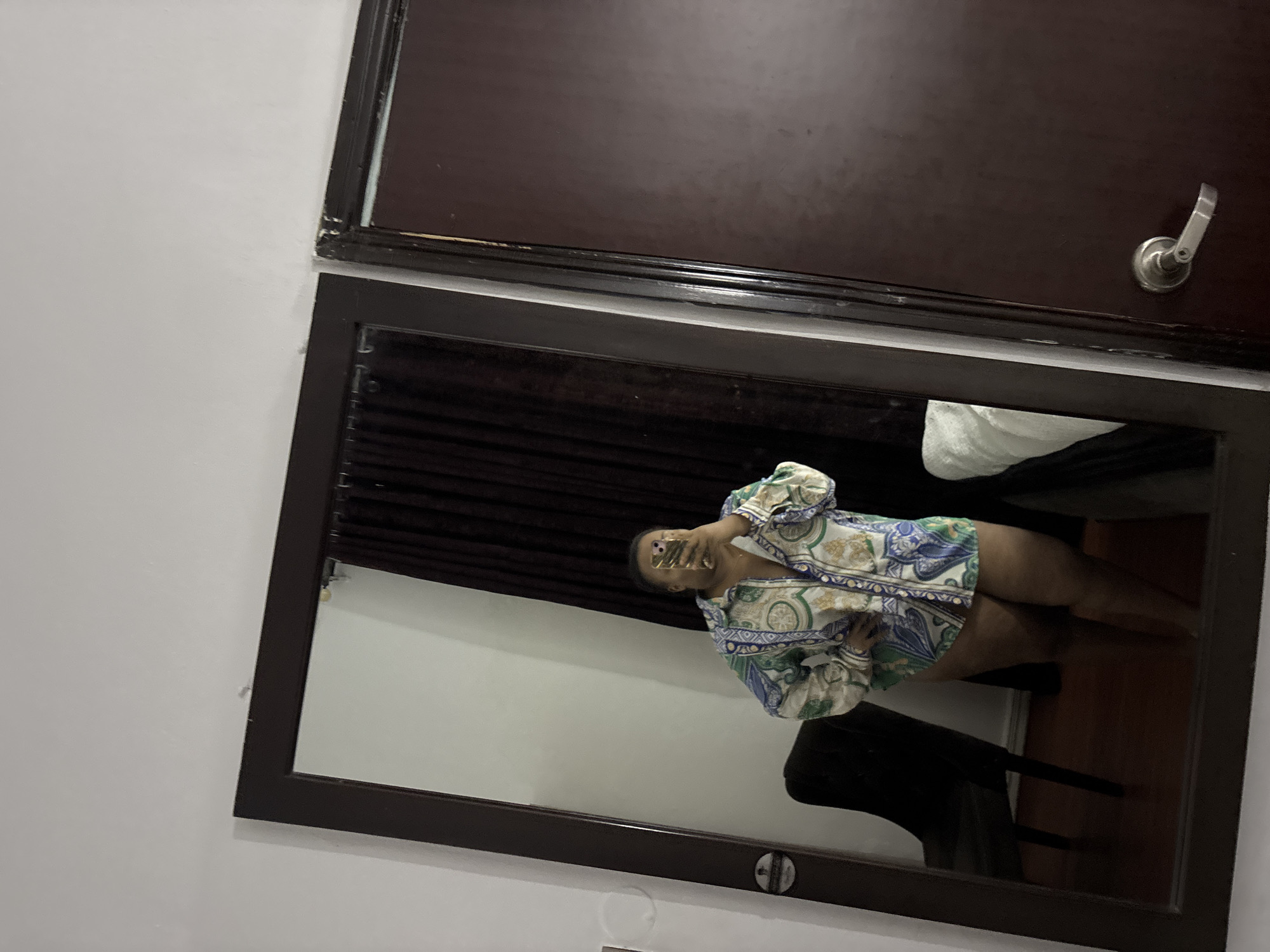 A mirror reflects a woman wearing a patterned shirt, appearing to take a selfie. Behind her, a dark curtain is visible. The setting includes a wooden door above the mirror and a chair within the room's reflection. It is not possible to determine if the location is in Port Harcourt, Nigeria from the image.
