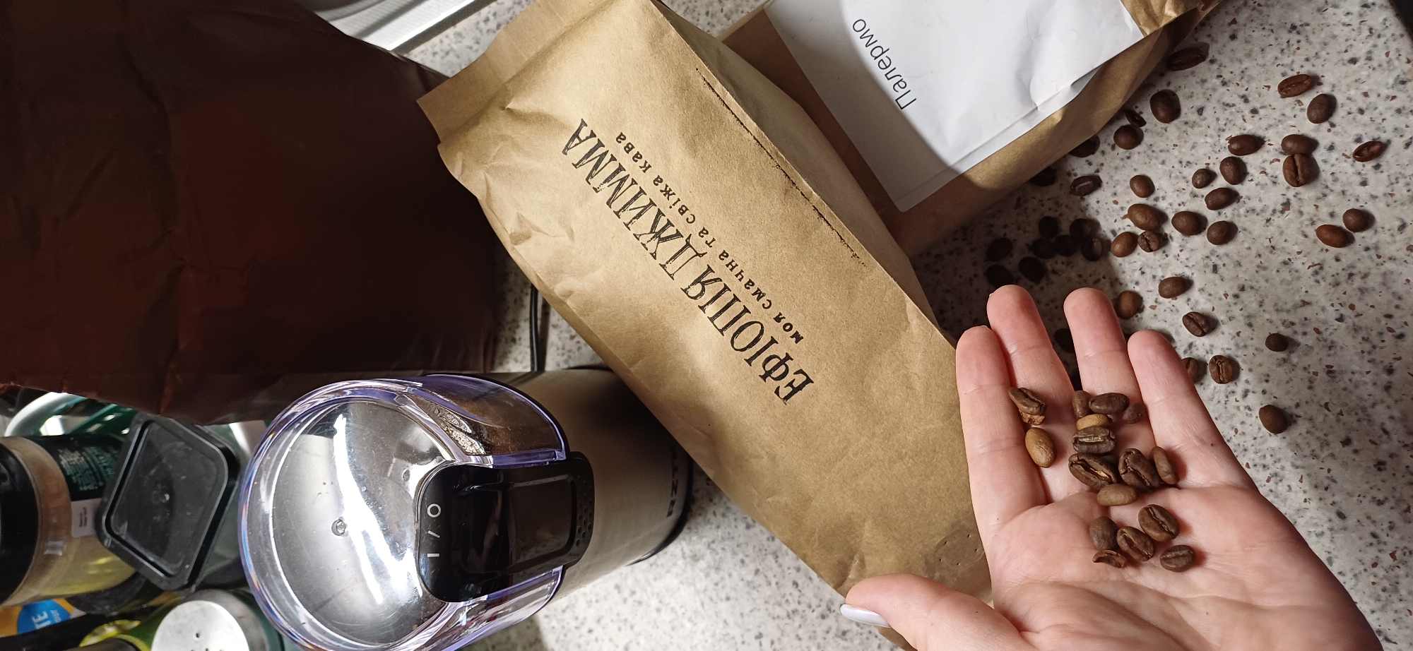 In an indoor setting, possibly a kitchen, a close-up shot focuses on various coffee-related items. In the foreground, a person's left hand, with a light-colored fingernail, holds a small pile of roasted coffee beans. The hand is positioned over a speckled gray countertop where numerous coffee beans are scattered. Behind the hand, a brown paper bag with Ukrainian text, "ЕФІОПІЯ ДЖІММА" and "моя смачна та свіжа кава," is prominently displayed. To the right of this bag, another brown paper bag with a white label reading "Палермо" is partially visible. In the background, a clear plastic-lidded coffee grinder, containing ground coffee, sits on the countertop. Several other containers and a dark brown paper bag are also present in the background, creating a still-life arrangement of coffee-related products.